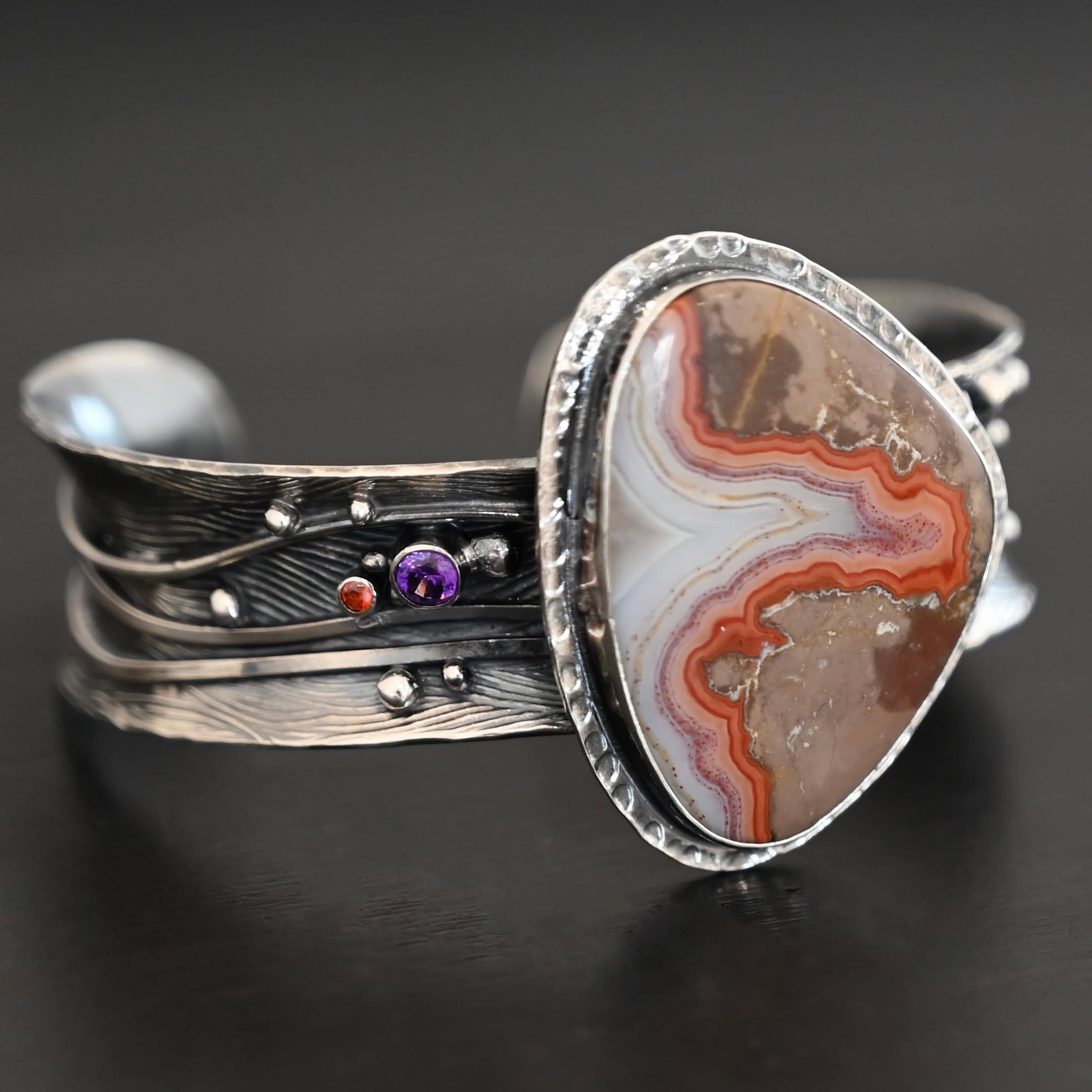 Zion Canyon Cuff "Zion Canyon" Cuff. There is a large, wide, oval, vertical ocean jasper in the center of a very wide, thick cuff. There are wavy lines of silver going across it and multiple gemstones scattered across it, along with silver ball detail. There is hammered silver around the stone.