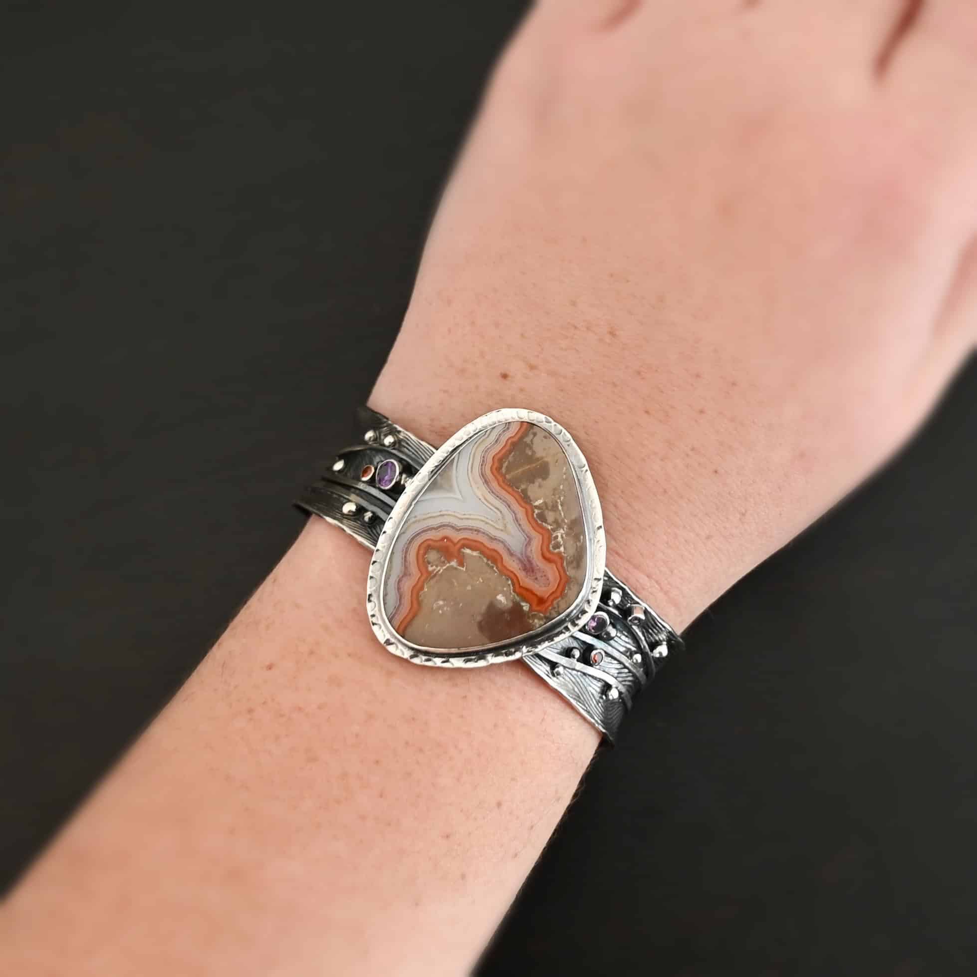 Zion Canyon Cuff "Zion Canyon" Cuff. There is a large, wide, oval, vertical ocean jasper in the center of a very wide, thick cuff. There are wavy lines of silver going across it and multiple gemstones scattered across it, along with silver ball detail. There is hammered silver around the stone. Necklace is shown up close by a model.