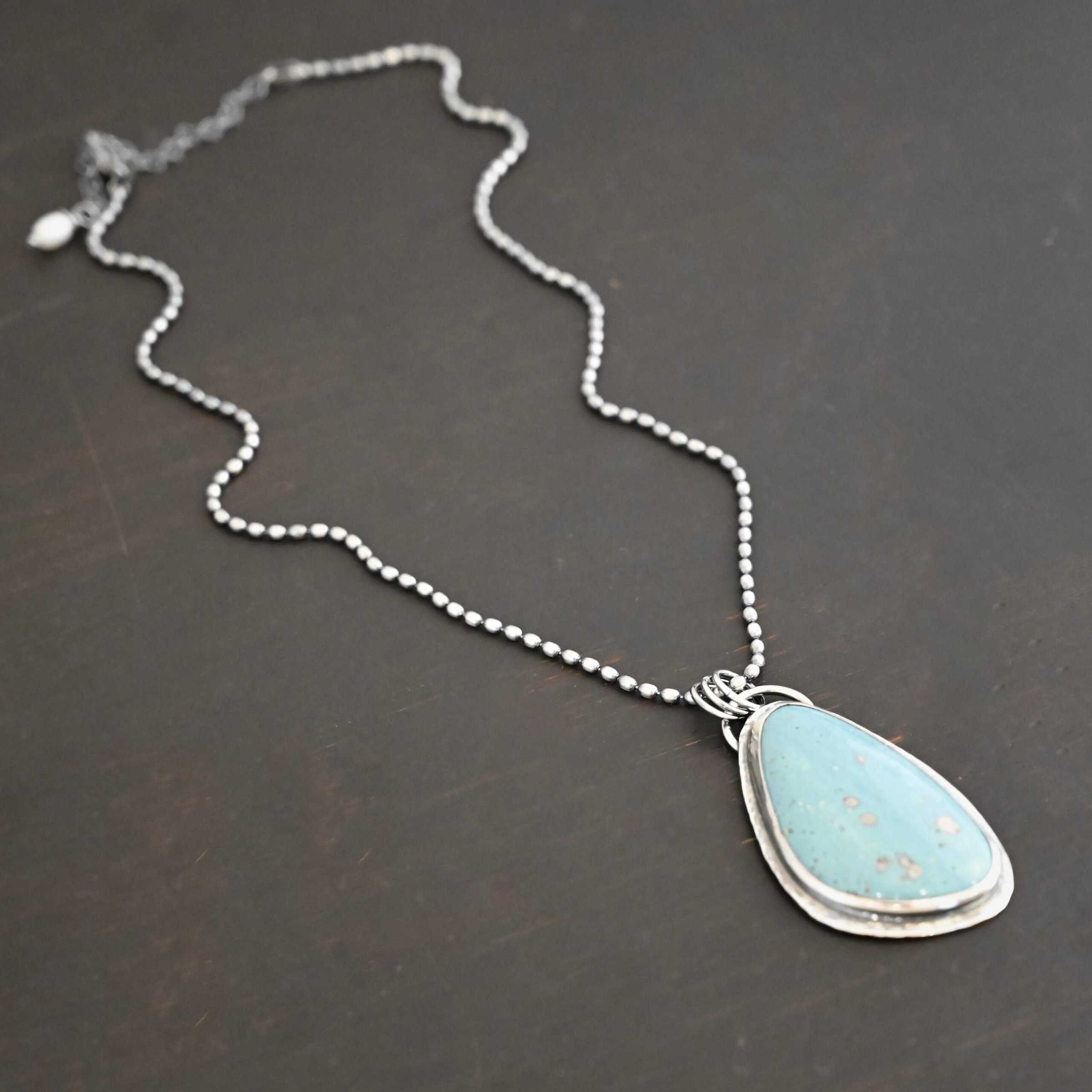 Close up of the "Galia" Necklace. Made out of sterling silver and leland blue. The leland blue is a light blue oval shape that is set vertically. There are some light gray dots scattered throughout the stone. There is a loop on the back of the pendant in which there are jump rings attached to it that are also attached to the chain of the necklace. The pendant is laying against a dark background.