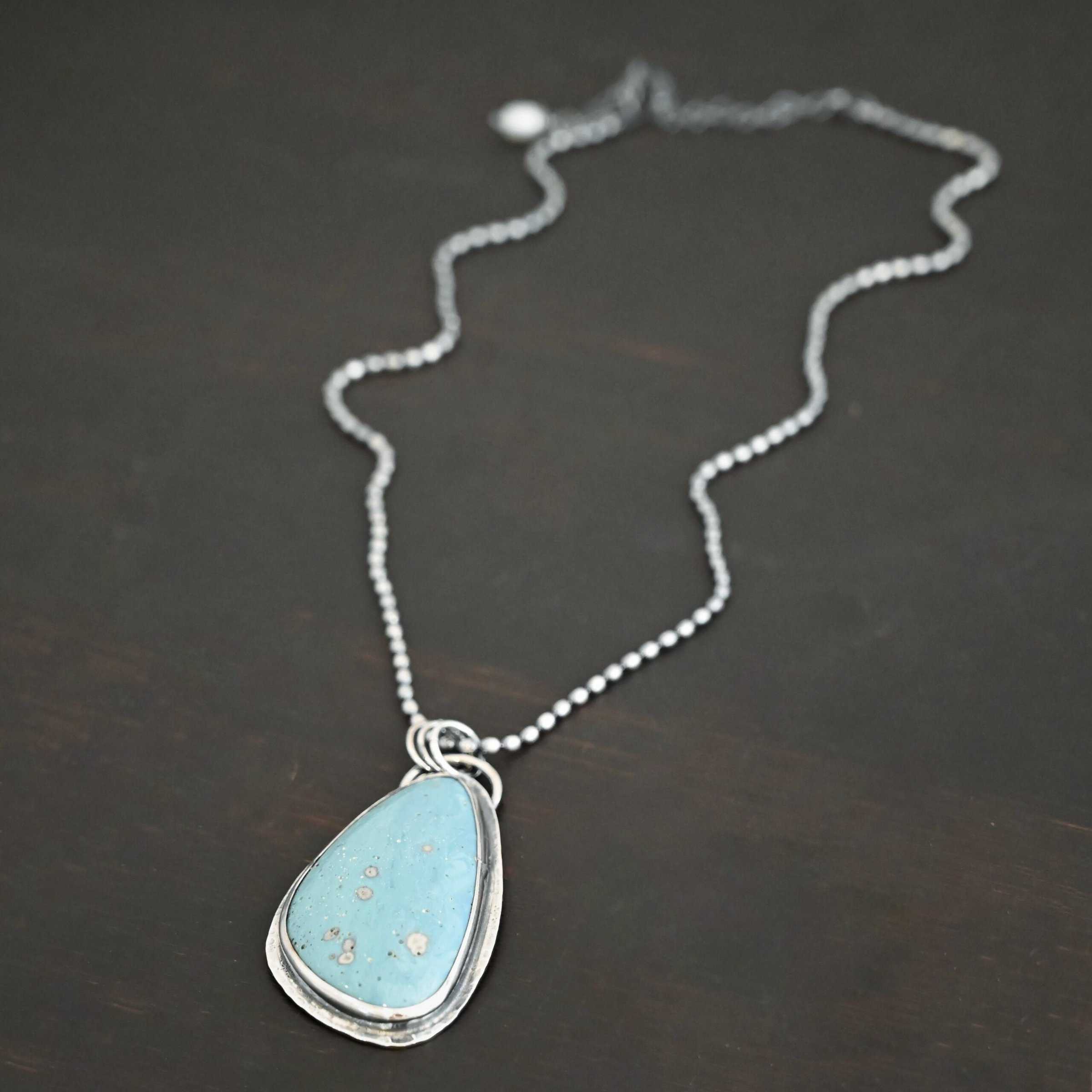 Close up of the "Galia" Necklace. Made out of sterling silver and leland blue. The leland blue is a light blue oval shape that is set vertically. There are some light gray dots scattered throughout the stone. There is a loop on the back of the pendant in which there are jump rings attached to it that are also attached to the chain of the necklace. The pendant is laying against a dark background.