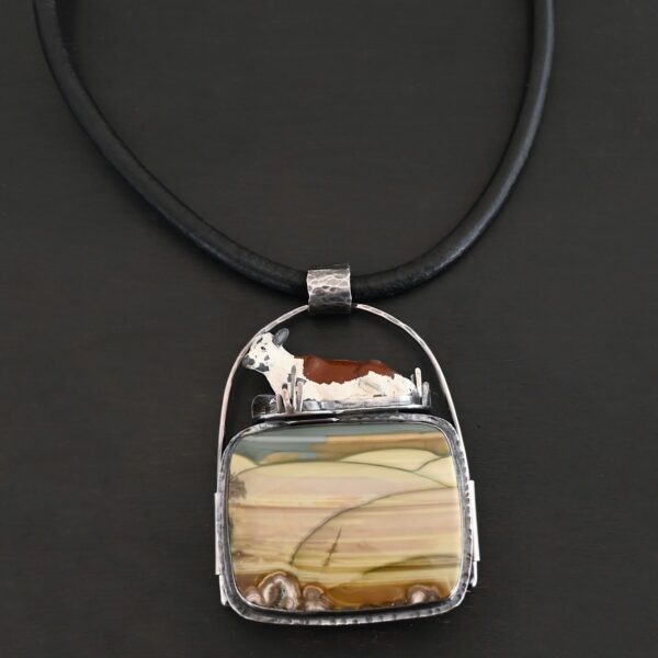 Close up of the "Till The Cows Come Home" Necklace. Made out of sterling silver and Morrisonite Jasper. The morrisonite jasper is a square shape, and is a mix of green and brown colors. There are dark green lines that are spread across the necklace to make it look as if there is a couple of grass hills. There is a small toy metal cow that is laying on top of the necklace. It is brown and white. The pendant has a large loop going across the back of it that is attached to a thick silver circle that is attached to a black leather chain. Necklace is laying against a black background.