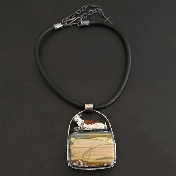 Close up of the "Till The Cows Come Home" Necklace. Made out of sterling silver and Morrisonite Jasper. The morrisonite jasper is a square shape, and is a mix of green and brown colors. There are dark green lines that are spread across the necklace to make it look as if there is a couple of grass hills. There is a small toy metal cow that is laying on top of the necklace. It is brown and white. The pendant has a large loop going across the back of it that is attached to a thick silver circle that is attached to a black leather chain. Necklace is laying against a black background.