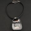 Close up of the "Till The Cows Come Home" Necklace. This shows the back of the pendant. There is a small silver cowbell that jingles when moved. There are also two small silver engraved wheat stalks on it. Also, there is the phrase "Until the cows come home" that is engraved into the back. There is a small plate on the top of the back that has "Wild Prairie Silver" hammered into it. Pendant is set against a dark background.