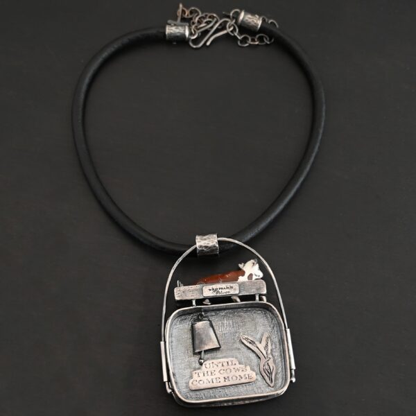 Close up of the "Till The Cows Come Home" Necklace. This shows the back of the pendant. There is a small silver cowbell that jingles when moved. There are also two small silver engraved wheat stalks on it. Also, there is the phrase "Until the cows come home" that is engraved into the back. There is a small plate on the top of the back that has "Wild Prairie Silver" hammered into it. Pendant is set against a dark background.