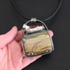 Close up of the "Till The Cows Come Home" Necklace. Made out of sterling silver and Morrisonite Jasper. The morrisonite jasper is a square shape, and is a mix of green and brown colors. There are dark green lines that are spread across the necklace to make it look as if there is a couple of grass hills. There is a small toy metal cow that is laying on top of the necklace. It is brown and white. The pendant has a large loop going across the back of it that is attached to a thick silver circle that is attached to a black leather chain. Necklace is laying against the palm of a hand to demonstrate size.