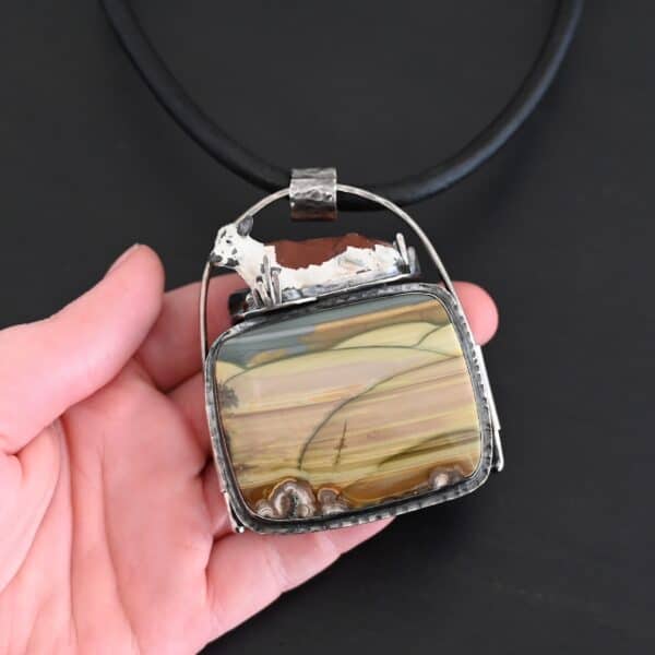Close up of the "Till The Cows Come Home" Necklace. Made out of sterling silver and Morrisonite Jasper. The morrisonite jasper is a square shape, and is a mix of green and brown colors. There are dark green lines that are spread across the necklace to make it look as if there is a couple of grass hills. There is a small toy metal cow that is laying on top of the necklace. It is brown and white. The pendant has a large loop going across the back of it that is attached to a thick silver circle that is attached to a black leather chain. Necklace is laying against the palm of a hand to demonstrate size.