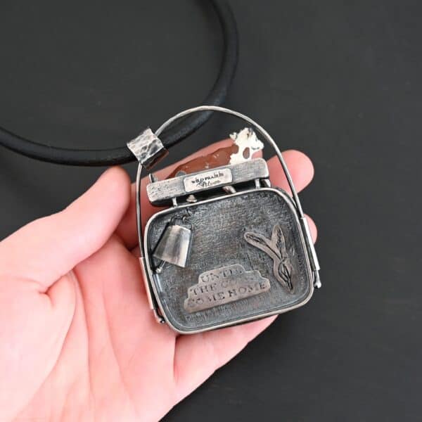 Close up of the "Till The Cows Come Home" Necklace. This shows the back of the pendant. There is a small silver cowbell that jingles when moved. There are also two small silver engraved wheat stalks on it. Also, there is the phrase "Until the cows come home" that is engraved into the back. There is a small plate on the top of the back that has "Wild Prairie Silver" hammered into it. Pendant is set against the palm of a hand to demonstrate size.
