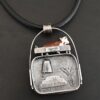 Close up of the "Till The Cows Come Home" Necklace. This shows the back of the pendant. There is a small silver cowbell that jingles when moved. There are also two small silver engraved wheat stalks on it. Also, there is the phrase "Until the cows come home" that is engraved into the back. There is a small plate on the top of the back that has "Wild Prairie Silver" hammered into it. Pendant is set against a dark background.