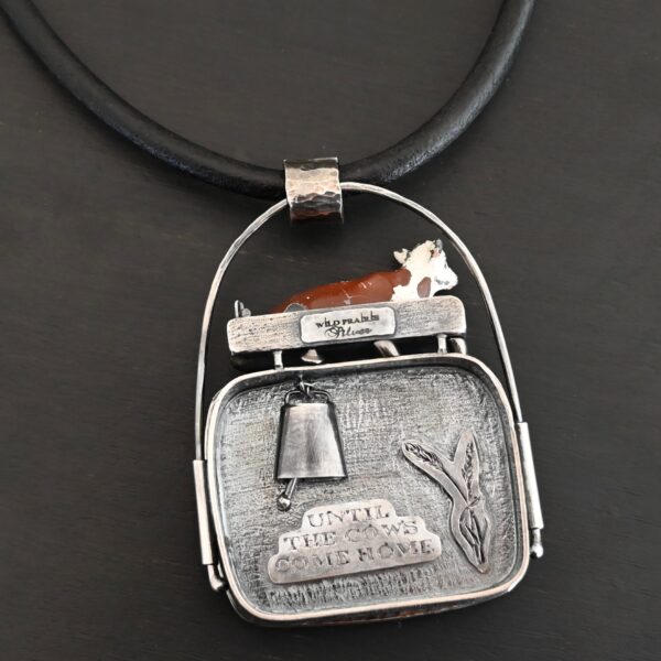Close up of the "Till The Cows Come Home" Necklace. This shows the back of the pendant. There is a small silver cowbell that jingles when moved. There are also two small silver engraved wheat stalks on it. Also, there is the phrase "Until the cows come home" that is engraved into the back. There is a small plate on the top of the back that has "Wild Prairie Silver" hammered into it. Pendant is set against a dark background.