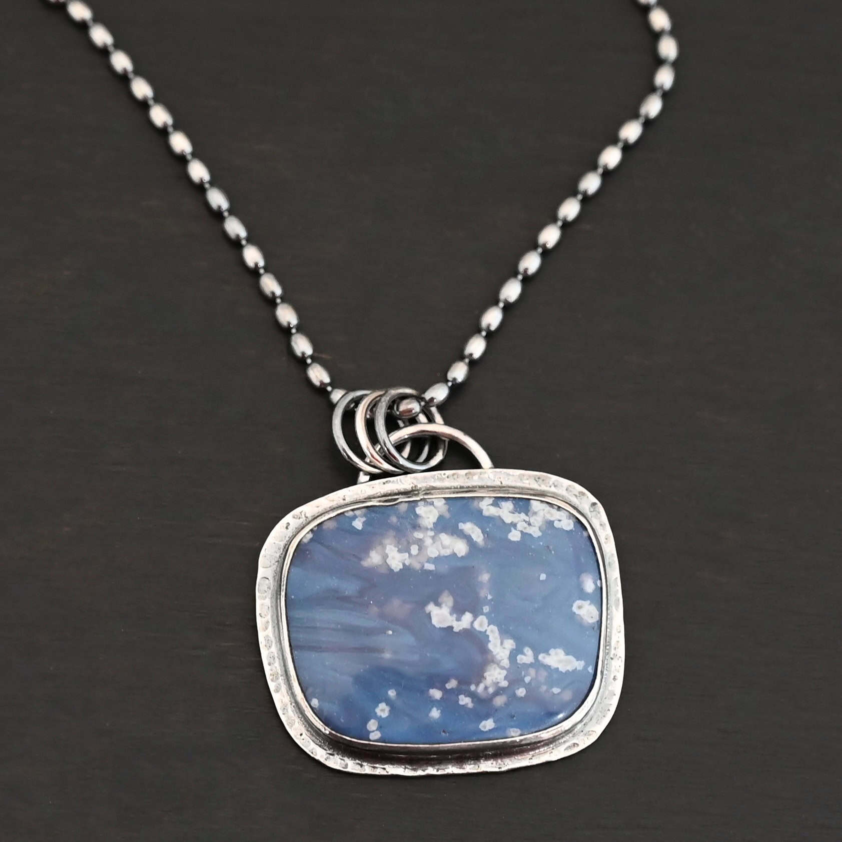 Close up of the "Azula" Necklace. Made out of sterling silver and leland blue. The leland blue is a square shape and is a dark blue color with light gray dots scattered throughout the stone. There is a loop on the back of the pendant that is attached to jump rings that are attached to the chain of the necklace. Pendant is laying against a dark background.