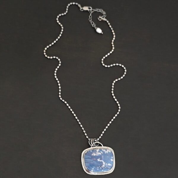 Close up of the "Azula" Necklace. Made out of sterling silver and leland blue. The leland blue is a square shape and is a dark blue color with light gray dots scattered throughout the stone. There is a loop on the back of the pendant that is attached to jump rings that are attached to the chain of the necklace. Pendant is laying against a dark background.