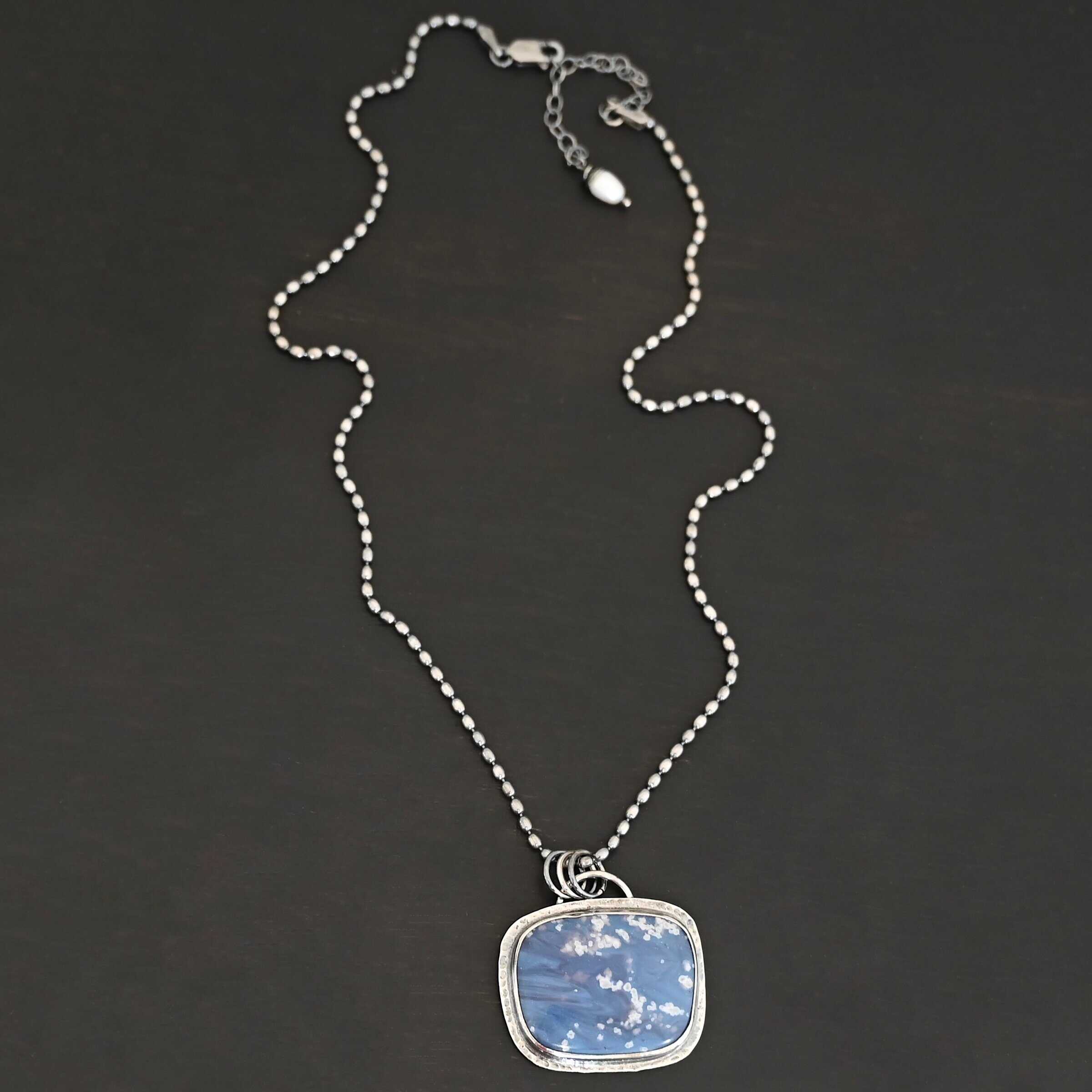Close up of the "Azula" Necklace. Made out of sterling silver and leland blue. The leland blue is a square shape and is a dark blue color with light gray dots scattered throughout the stone. There is a loop on the back of the pendant that is attached to jump rings that are attached to the chain of the necklace. Pendant is laying against a dark background.