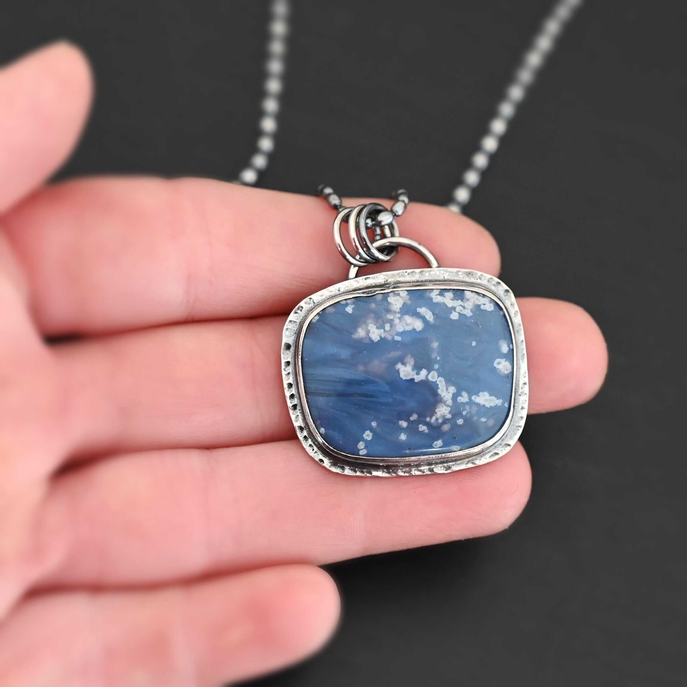 Close up of the "Azula" Necklace. Made out of sterling silver and leland blue. The leland blue is a square shape and is a dark blue color with light gray dots scattered throughout the stone. There is a loop on the back of the pendant that is attached to jump rings that are attached to the chain of the necklace. Pendant is laying on the fingertips of a hand to demonstrate size.