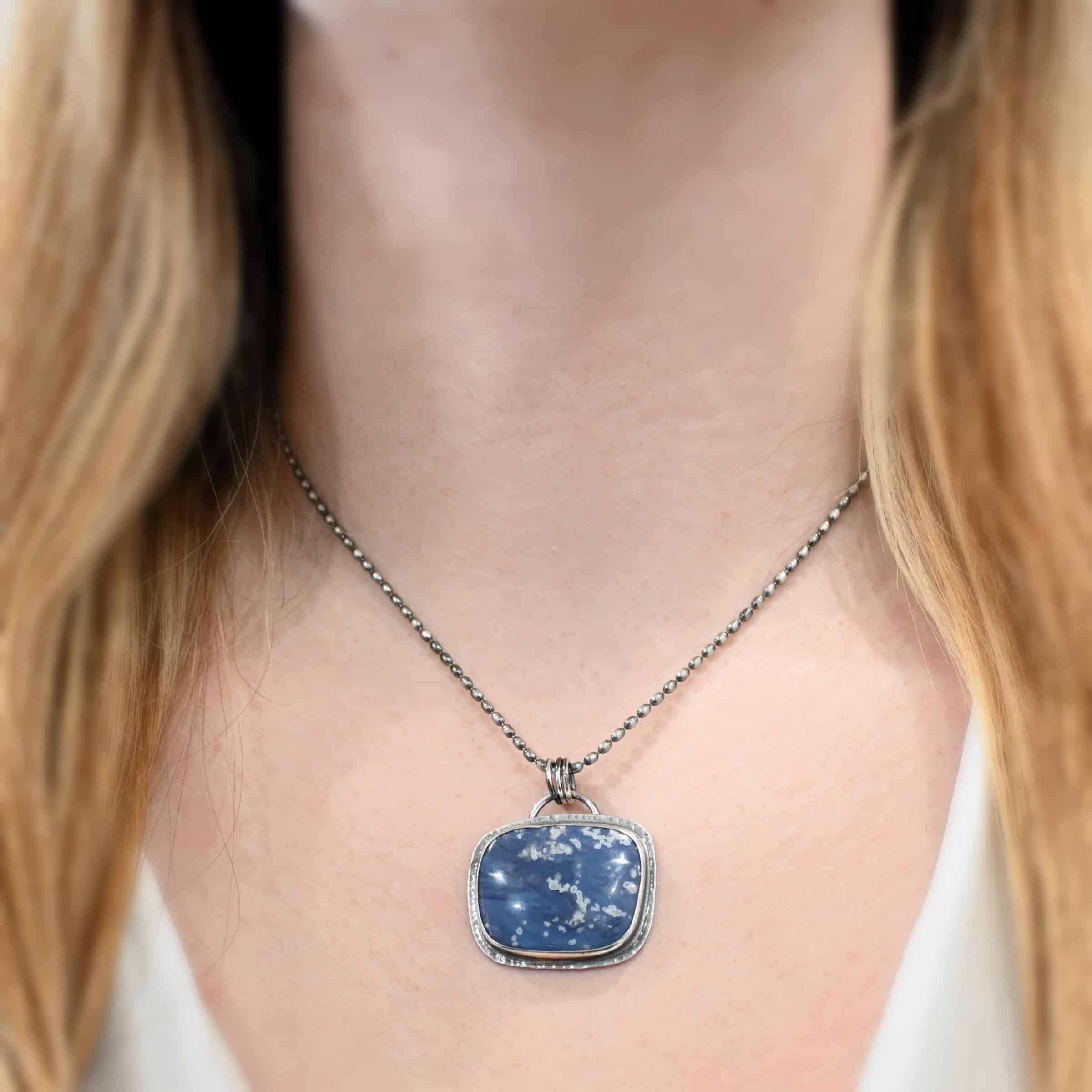 Close up of the "Azula" Necklace. Made out of sterling silver and leland blue. The leland blue is a square shape and is a dark blue color with light gray dots scattered throughout the stone. There is a loop on the back of the pendant that is attached to jump rings that are attached to the chain of the necklace. Pendant is worn by a model at 18 inches long to demonstrate length. Model is wearing a white v-neck shirt.