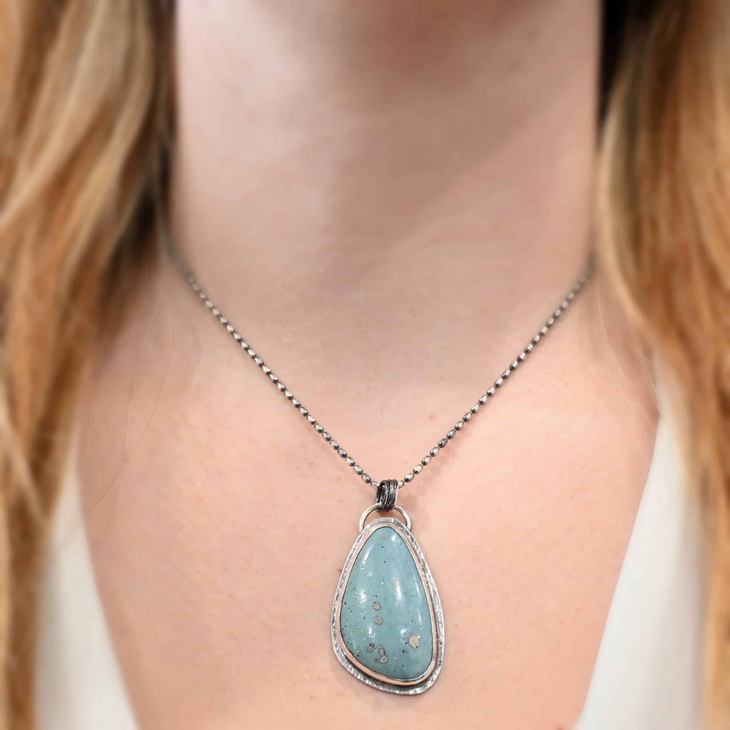 Close up of the "Galia" Necklace. Made out of sterling silver and leland blue. The leland blue is a light blue oval shape that is set vertically. There are some light gray dots scattered throughout the stone. There is a loop on the back of the pendant in which there are jump rings attached to it that are also attached to the chain of the necklace. Pendant is worn by a model at 18 inches long who is wearing a white v-neck shirt.