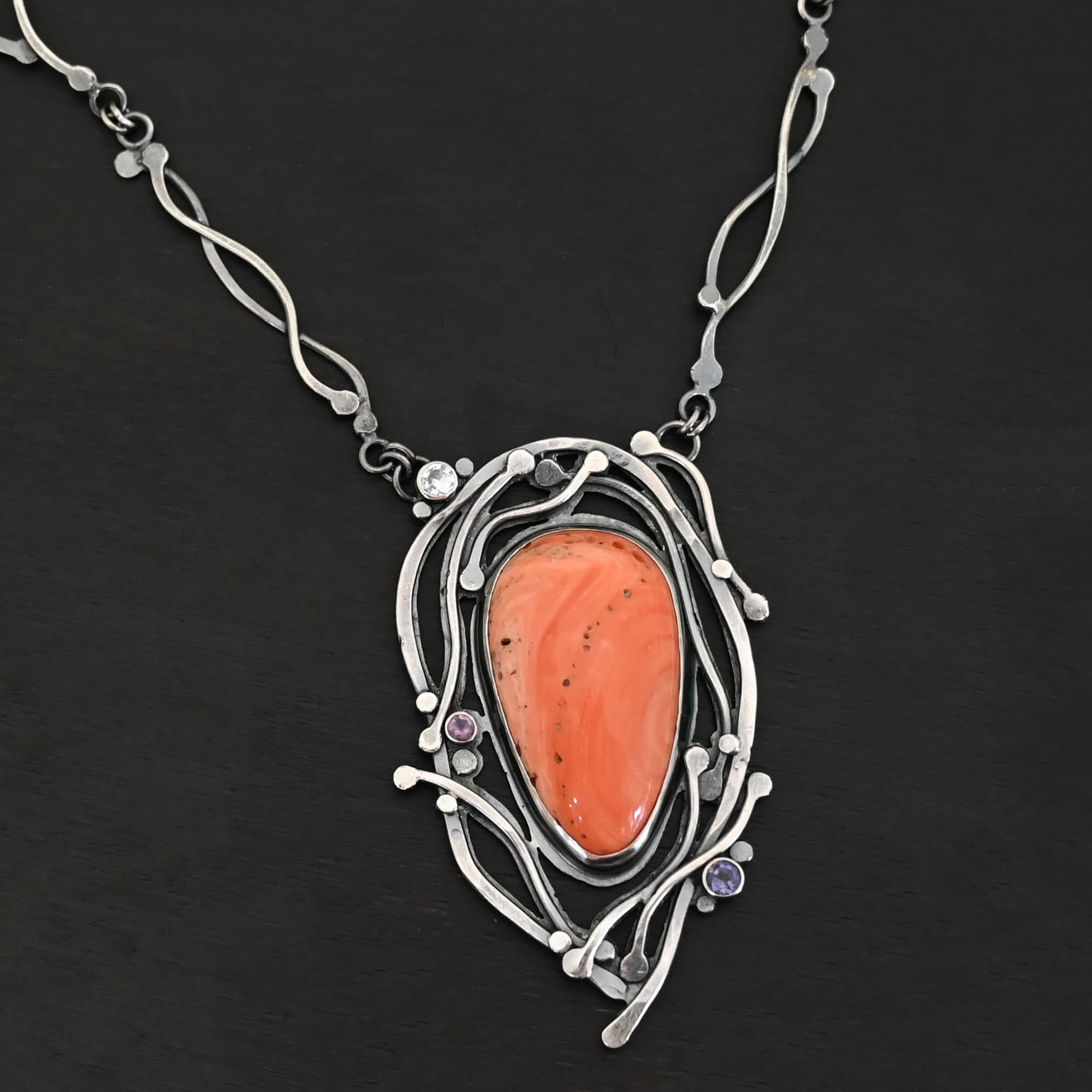 Hawaiian Breeze Necklace New Close up of the "Hawaiian Breeze" Necklace. Made out of sterling silver, palm coral fossil, and amethyst and aquamarine. The palm coral fossil is the biggest stone. It is an oval shape and a light orange color. There are small holes scattered throughout the necklace. There are two amethyst stones that are small and round on the necklace. One is in the center left side of the necklace attached to the fossil. It is a light purple. The other is on the right bottom side of the necklace. This one is a dark purple. The aquamarine is in the top left corner of the necklace. It is a very light blue. The rest of the necklace is surrounded by silver swirls overlapping one another. The pendant is attached on both sides at the top with jump rings to the chain. Necklace is set against a dark background.