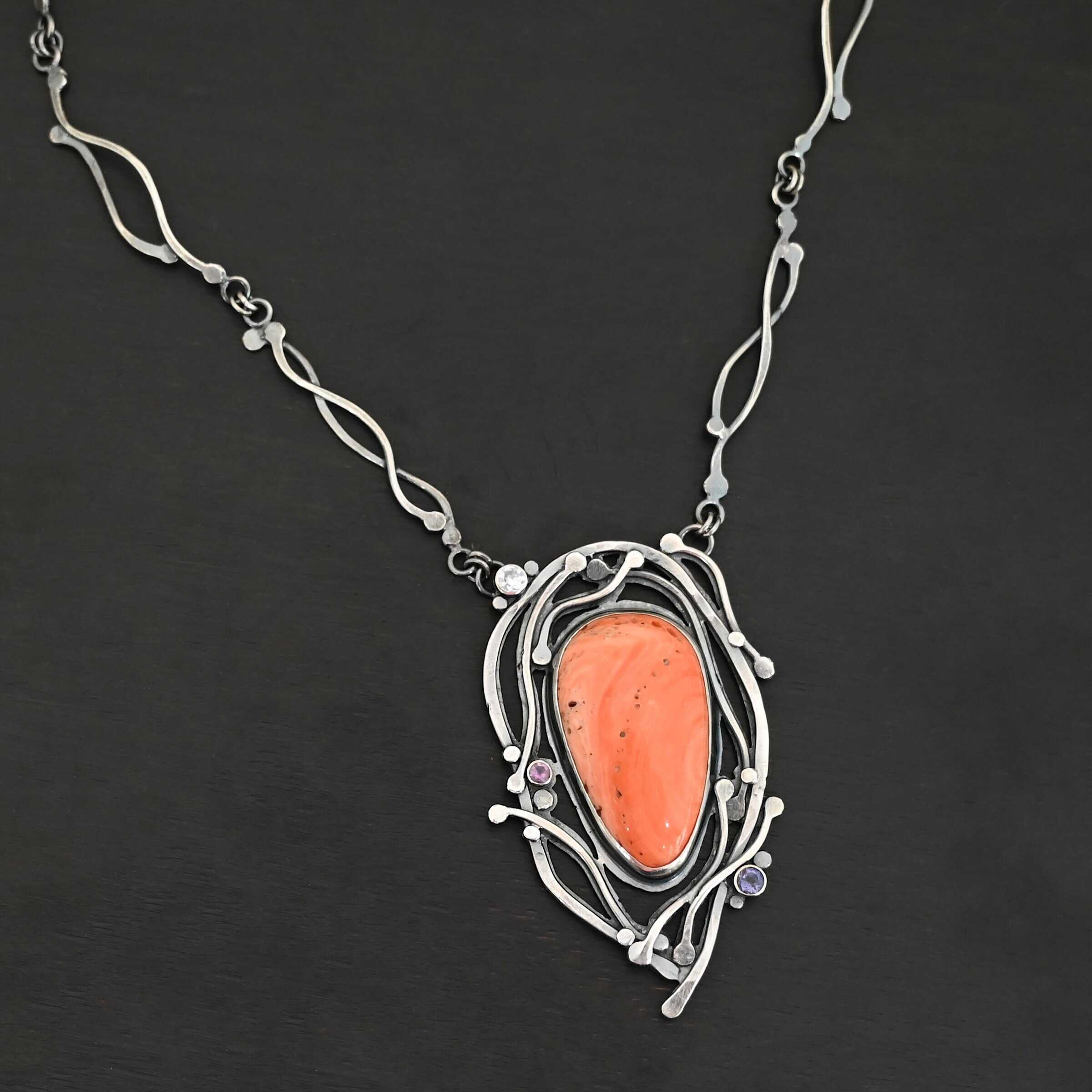 Hawaiian Breeze Necklace New Close up of the "Hawaiian Breeze" Necklace. Made out of sterling silver, palm coral fossil, and amethyst and aquamarine. The palm coral fossil is the biggest stone. It is an oval shape and a light orange color. There are small holes scattered throughout the necklace. There are two amethyst stones that are small and round on the necklace. One is in the center left side of the necklace attached to the fossil. It is a light purple. The other is on the right bottom side of the necklace. This one is a dark purple. The aquamarine is in the top left corner of the necklace. It is a very light blue. The rest of the necklace is surrounded by silver swirls overlapping one another. The pendant is attached on both sides at the top with jump rings to the chain. Necklace is set against a dark background.