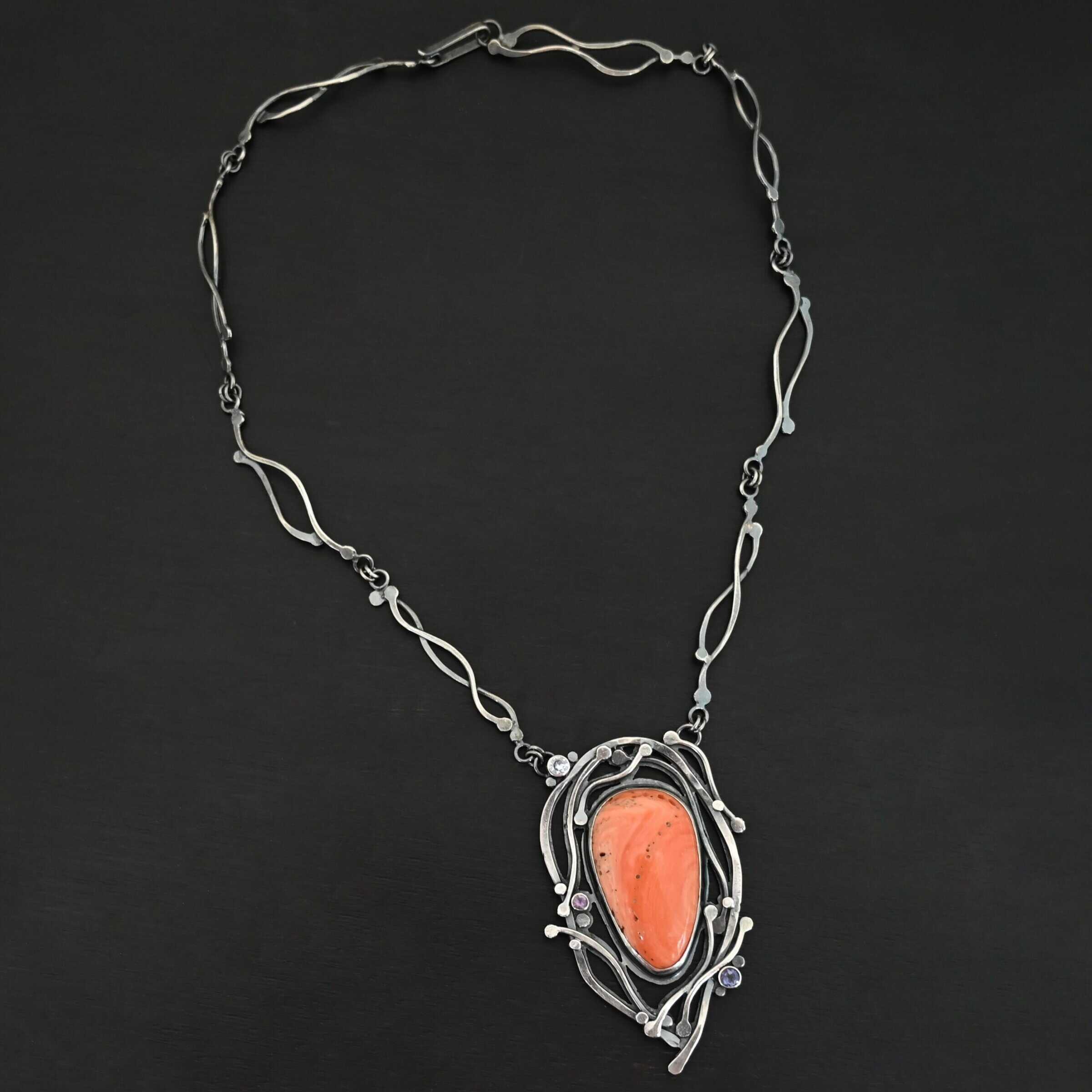 Hawaiian Breeze Necklace New Close up of the "Hawaiian Breeze" Necklace. Made out of sterling silver, palm coral fossil, and amethyst and aquamarine. The palm coral fossil is the biggest stone. It is an oval shape and a light orange color. There are small holes scattered throughout the necklace. There are two amethyst stones that are small and round on the necklace. One is in the center left side of the necklace attached to the fossil. It is a light purple. The other is on the right bottom side of the necklace. This one is a dark purple. The aquamarine is in the top left corner of the necklace. It is a very light blue. The rest of the necklace is surrounded by silver swirls overlapping one another. The pendant is attached on both sides at the top with jump rings to the chain. Necklace is set against a dark background.