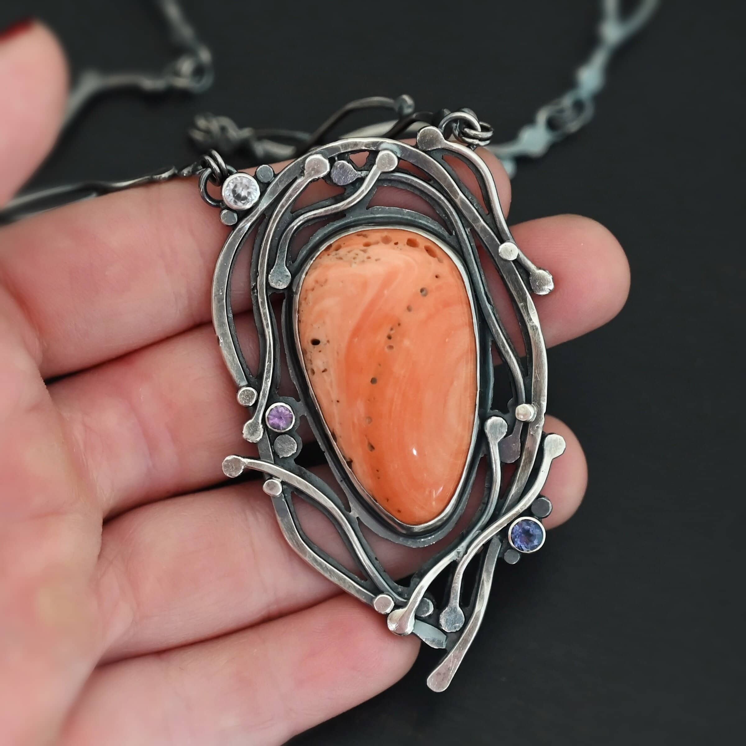 Hawaiian Breeze Necklace New Close up of the "Hawaiian Breeze" Necklace. Made out of sterling silver, palm coral fossil, and amethyst and aquamarine. The palm coral fossil is the biggest stone. It is an oval shape and a light orange color. There are small holes scattered throughout the necklace. There are two amethyst stones that are small and round on the necklace. One is in the center left side of the necklace attached to the fossil. It is a light purple. The other is on the right bottom side of the necklace. This one is a dark purple. The aquamarine is in the top left corner of the necklace. It is a very light blue. The rest of the necklace is surrounded by silver swirls overlapping one another. The pendant is attached on both sides at the top with jump rings to the chain. Necklace is set against the fingertips of a hand.