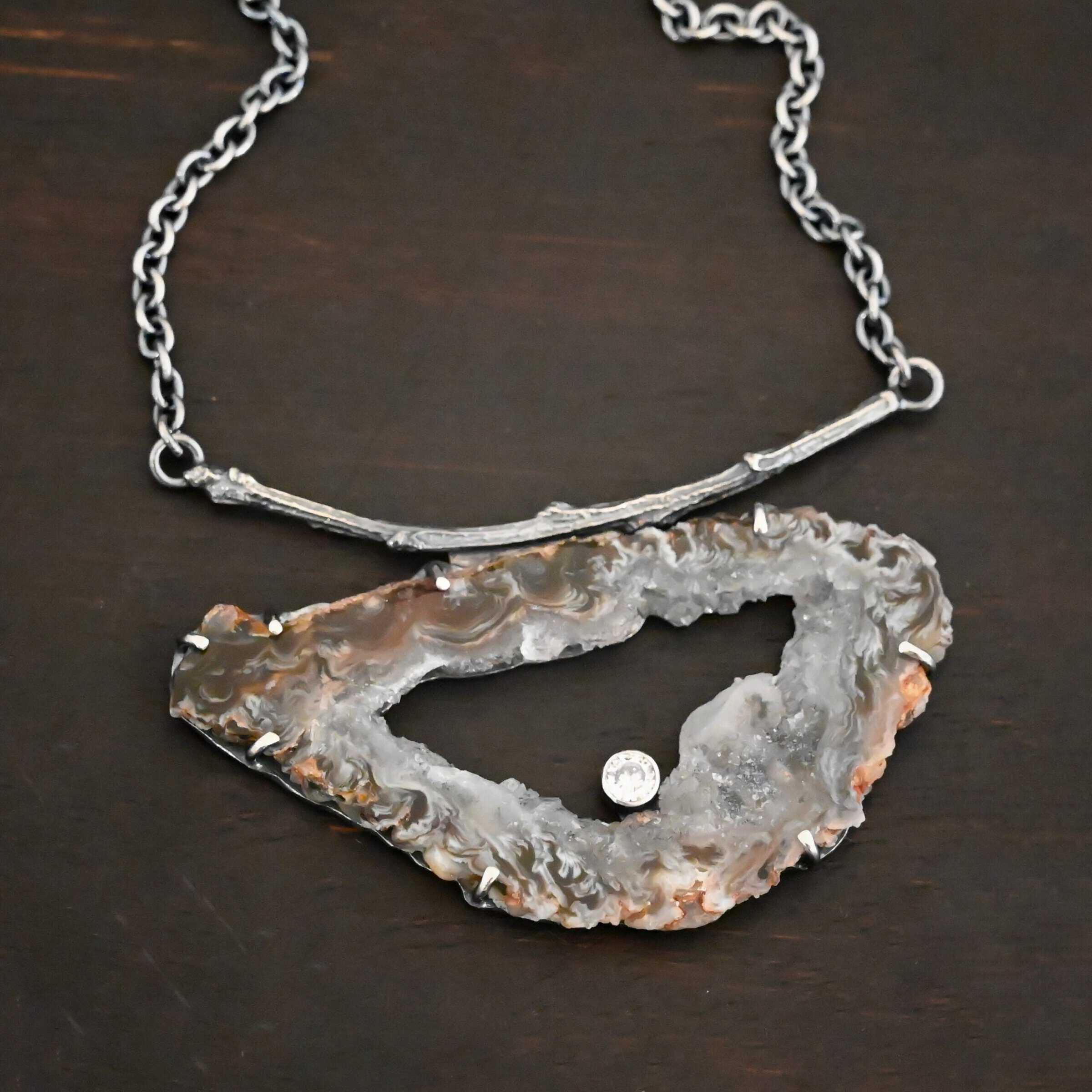 Close up of the "Carina" Necklace. Made out of sterling silver, druzy agate, and cubic zirconia. The druzy agate is the main stone, and it is mostly gray with some red swirls. There is a hole in the center of the stone that is a long oval shape, but slightly jagged. The cubic zirconia is in the center of the necklace in the hole and it is on the bottom of the ovular hole. The druzy is soldered to a casted sterling silver twig that is slightly curved. On both ends of the twig there are two loops that are connected to a chain that connects at the back to wear the necklace. Pendant is laying against a dark background.