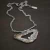 Close up of the "Carina" Necklace. Made out of sterling silver, druzy agate, and cubic zirconia. The druzy agate is the main stone, and it is mostly gray with some red swirls. There is a hole in the center of the stone that is a long oval shape, but slightly jagged. The cubic zirconia is in the center of the necklace in the hole and it is on the bottom of the ovular hole. The druzy is soldered to a casted sterling silver twig that is slightly curved. On both ends of the twig there are two loops that are connected to a chain that connects at the back to wear the necklace. Pendant is laying against a dark background.