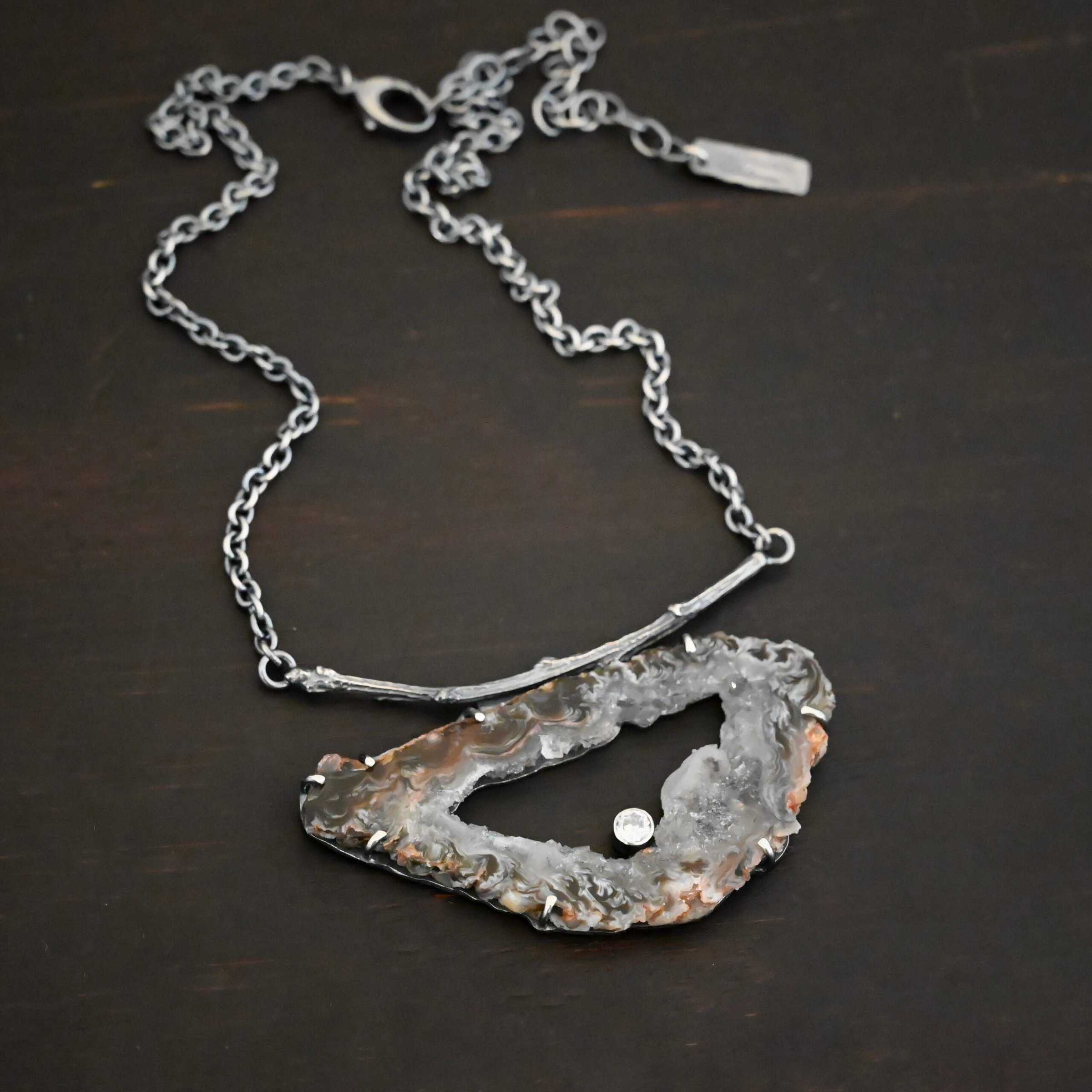 Close up of the "Carina" Necklace. Made out of sterling silver, druzy agate, and cubic zirconia. The druzy agate is the main stone, and it is mostly gray with some red swirls. There is a hole in the center of the stone that is a long oval shape, but slightly jagged. The cubic zirconia is in the center of the necklace in the hole and it is on the bottom of the ovular hole. The druzy is soldered to a casted sterling silver twig that is slightly curved. On both ends of the twig there are two loops that are connected to a chain that connects at the back to wear the necklace. Pendant is laying against a dark background.