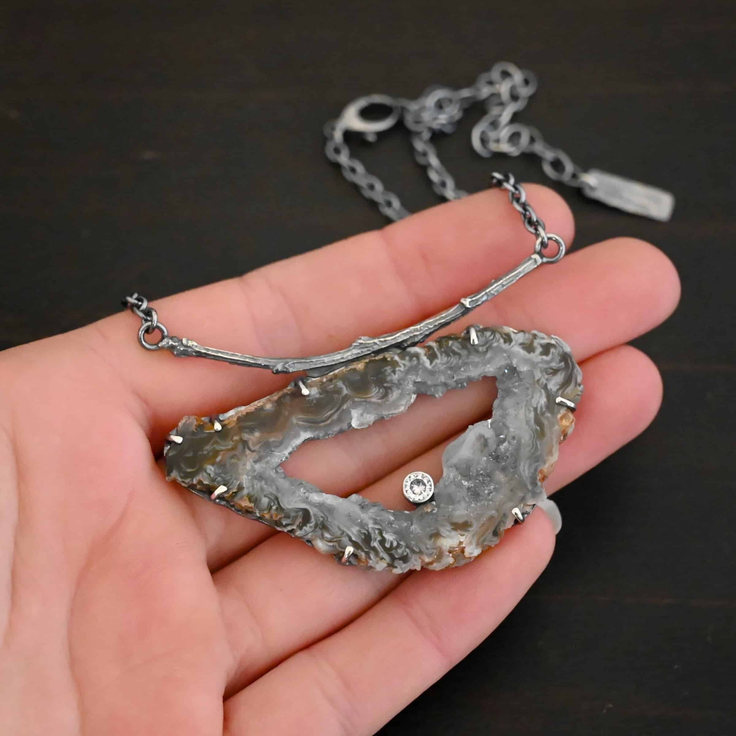 Close up of the "Carina" Necklace. Made out of sterling silver, druzy agate, and cubic zirconia. The druzy agate is the main stone, and it is mostly gray with some red swirls surrounding it. There is a hole in the center of the stone that is a long oval shape, but slightly jagged. The cubic zirconia is in the center of the necklace in the hole and it is on the bottom of the ovular hole. The druzy is soldered to a casted sterling silver twig that is slightly curved. On both ends of the twig there are two loops that are connected to a chain that connects at the back to wear the necklace. Necklace is laying against the fingertips of a hand to demonstrate size of the pendant.