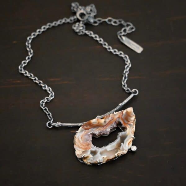 Close up of the "Astra" Necklace. Made out of sterling silver, druzy agate, and cubic zirconia. The druzy agate is the main stone, and it is a mix of red and brown swirls. There is a hole in the center of the stone that is a long oval shape, but slightly jagged. Surrounding the edges of the hole and going around it is a purple color. The cubic zirconia is on the bottom center of the necklace. The druzy is soldered to a casted sterling silver twig that is slightly curved. On both ends of the twig there are two loops that are connected to a chain that connects at the back to wear the necklace. Pendant is laying against a dark background.