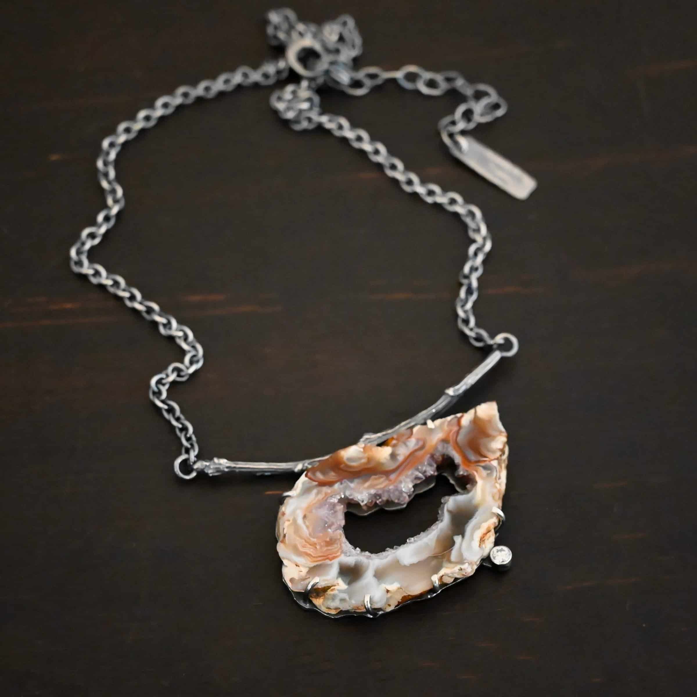 Astra Necklace Close up of the "Astra" Necklace. Made out of sterling silver, druzy agate, and cubic zirconia. The druzy agate is the main stone, and it is a mix of red and brown swirls. There is a hole in the center of the stone that is a long oval shape, but slightly jagged. Surrounding the edges of the hole and going around it is a purple color. The cubic zirconia is on the bottom center of the necklace. The druzy is soldered to a casted sterling silver twig that is slightly curved. On both ends of the twig there are two loops that are connected to a chain that connects at the back to wear the necklace. Pendant is laying against a dark background.