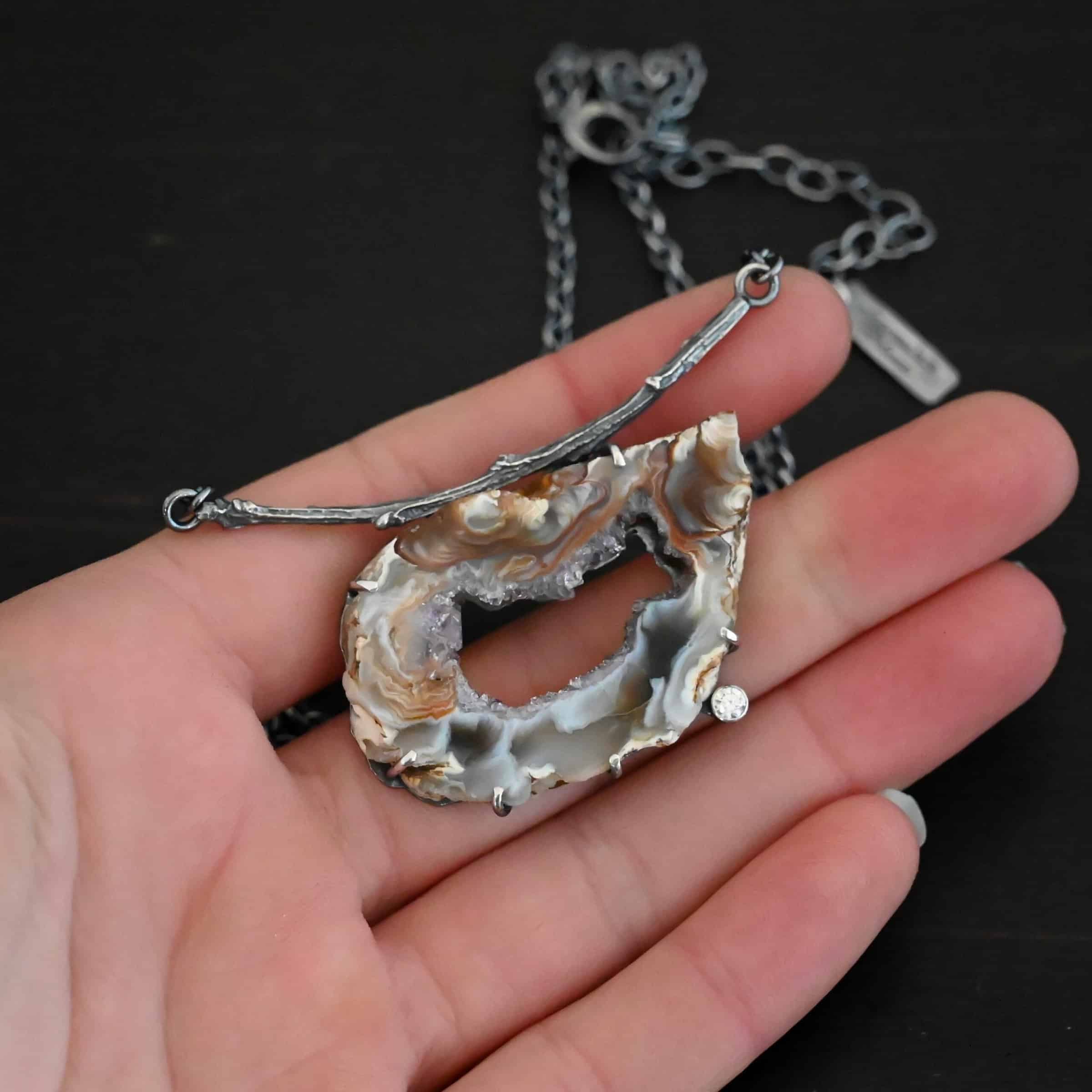 Astra Necklace Close up of the "Astra" Necklace. Made out of sterling silver, druzy agate, and cubic zirconia. The druzy agate is the main stone, and it is a mix of red and brown swirls. There is a hole in the center of the stone that is a long oval shape, but slightly jagged. Surrounding the edges of the hole and going around it is a purple color. The cubic zirconia is on the bottom center of the necklace. The druzy is soldered to a casted sterling silver twig that is slightly curved. On both ends of the twig there are two loops that are connected to a chain that connects at the back to wear the necklace. Pendant is laying against a dark background. Necklace is laying against the fingertips of a hand to demonstrate size of the pendant.