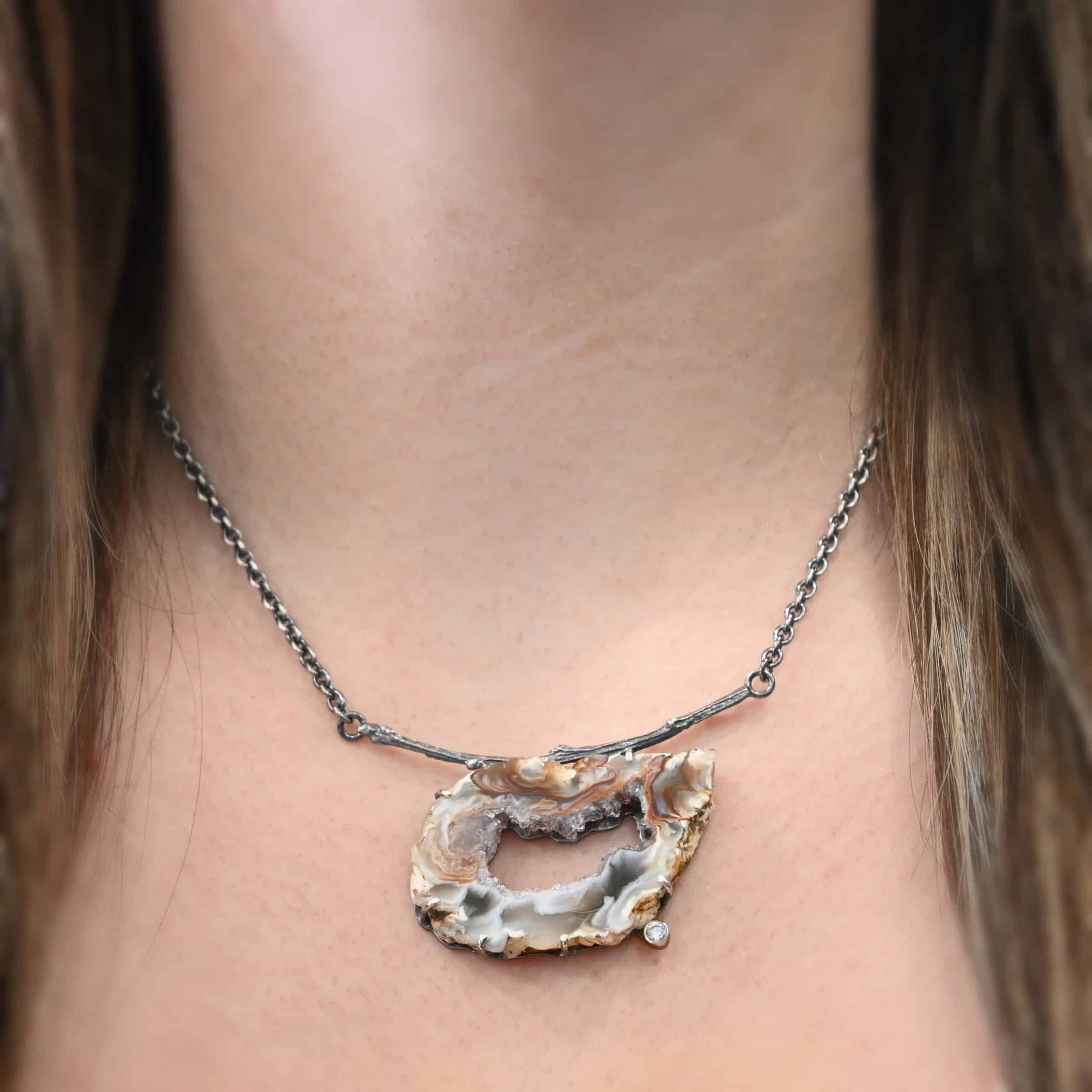 Astra Necklace Close up of the "Astra" Necklace on a model. Made out of sterling silver, druzy agate, and cubic zirconia. The druzy agate is the main stone, and it is a mix of red and brown swirls. There is a hole in the center of the stone that is a long oval shape, but slightly jagged. Surrounding the edges of the hole and going around it is a purple color. The cubic zirconia is on the bottom center of the necklace. The druzy is soldered to a casted sterling silver twig that is slightly curved. On both ends of the twig there are two loops that are connected to a chain that connects at the back to wear the necklace. Necklace is worn by a model.