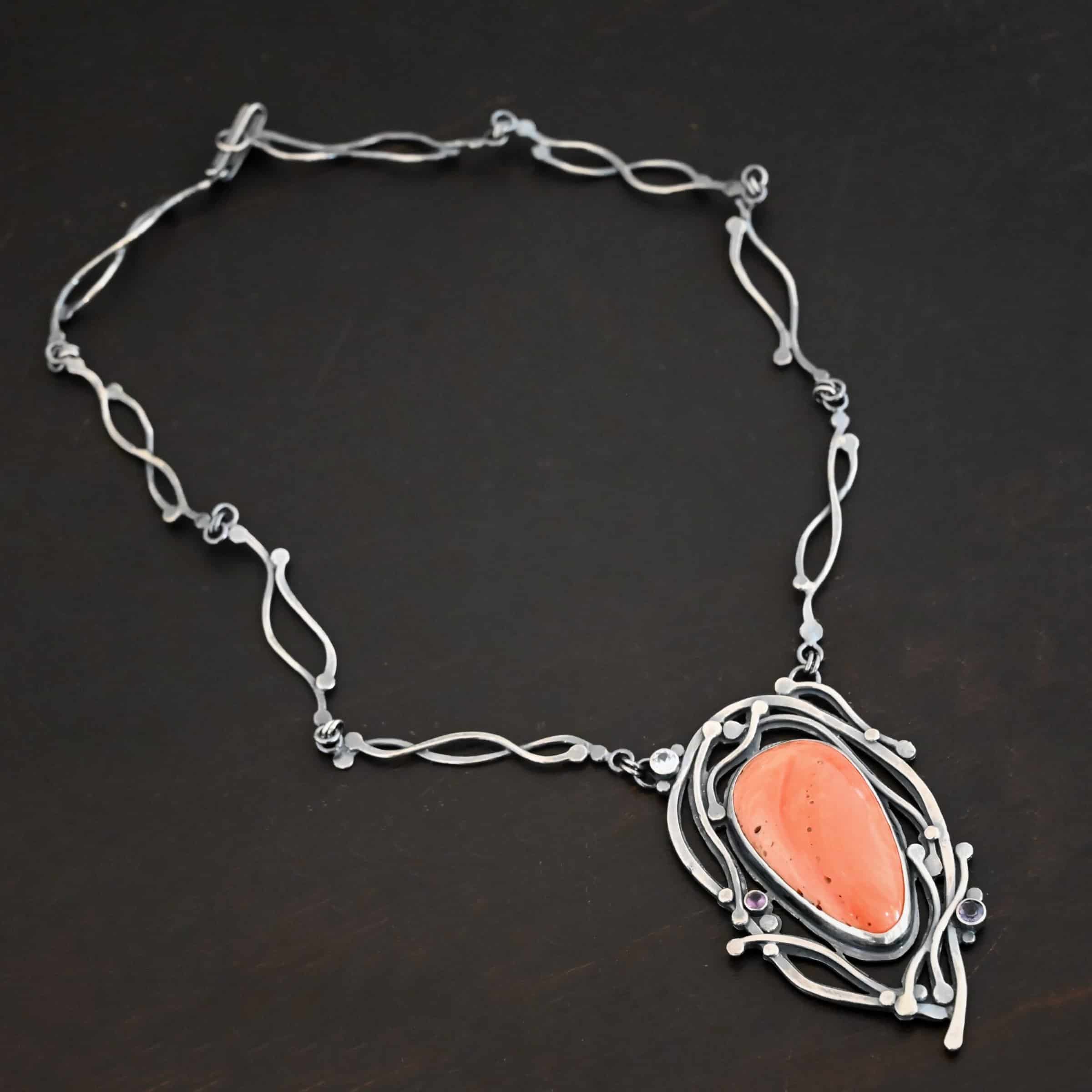 Hawaiian Breeze Necklace Close up of the "Hawaiian Breeze" Necklace. Made out of sterling silver, palm coral fossil, and amethyst and aquamarine. The palm coral fossil is the biggest stone. It is an oval shape and a light orange color. There are small holes scattered throughout the necklace. There are two amethyst stones that are small and round on the necklace. One is in the center left side of the necklace attached to the fossil. It is a light purple. The other is on the right bottom side of the necklace. This one is a dark purple. The aquamarine is in the top left corner of the necklace. It is a very light blue. The rest of the necklace is surrounded by silver swirls overlapping one another. The pendant is attached on both sides at the top with jump rings to the chain. Necklace is set against a dark background.