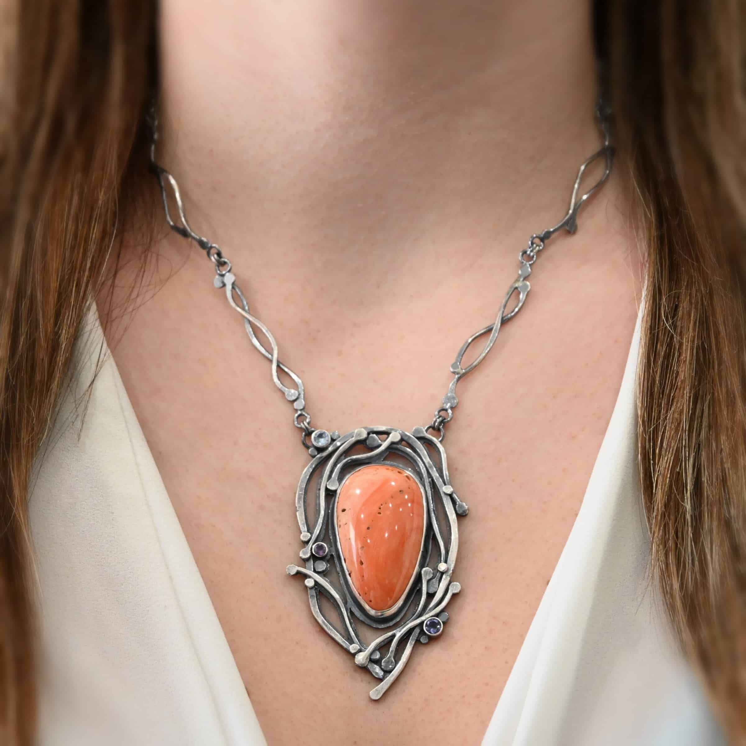 Hawaiian Breeze Necklace Close up of the "Hawaiian Breeze" Necklace. Made out of sterling silver, palm coral fossil, and amethyst and aquamarine. The palm coral fossil is the biggest stone. It is an oval shape and a light orange color. There are small holes scattered throughout the necklace. There are two amethyst stones that are small and round on the necklace. One is in the center left side of the necklace attached to the fossil. It is a light purple. The other is on the right bottom side of the necklace. This one is a dark purple. The aquamarine is in the top left corner of the necklace. It is a very light blue. The rest of the necklace is surrounded by silver swirls overlapping one another. The pendant is attached on both sides at the top with jump rings to the chain. Necklace is worn by a model to demonstrate size and length of the chain and pendant.