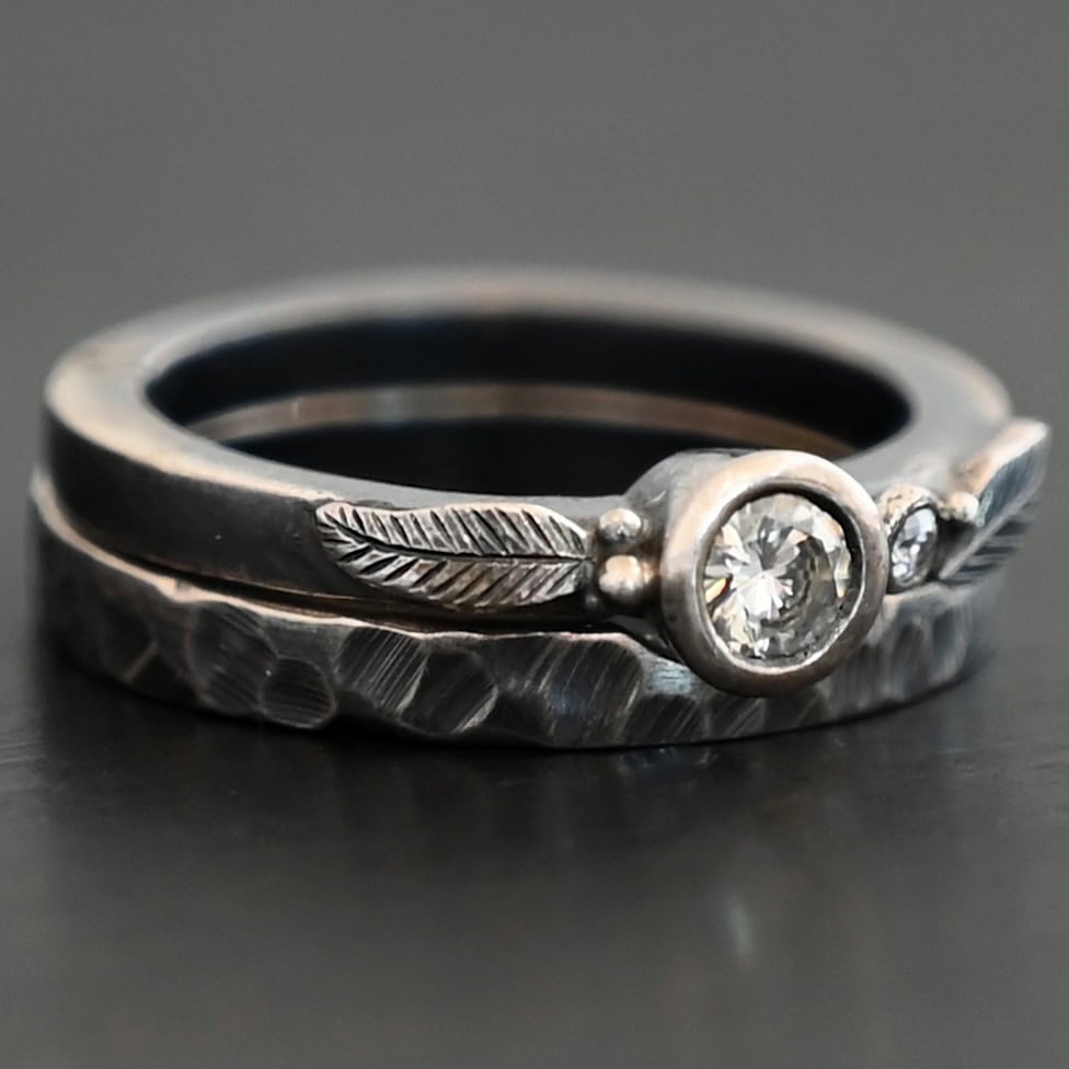 Close up of the "Shining Light" Ring. This ring is handmade out of sterling silver and moissanites. There are two moissanites ~ one as the center stone, and one on the right side of that. The ring has two separate bands that go together, as the top one sits on the bottom one in a groove. There are two silver leaves, two on the top band on each side of the center moissanite. The bottom band is hammered and oxidized silver. Ring is set against a dark background.