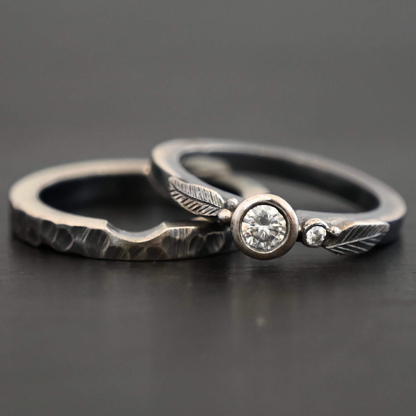 Close up of the "Shining Light" Ring. This ring is handmade out of sterling silver and moissanites. There are two moissanites ~ one as the center stone, and one on the right side of that. The ring has two separate bands that go together, as the top one sits on the bottom one in a groove. There are two silver leaves, two on the top band on each side of the center moissanite. The bottom band is hammered and oxidized silver. Ring is set against a dark background.