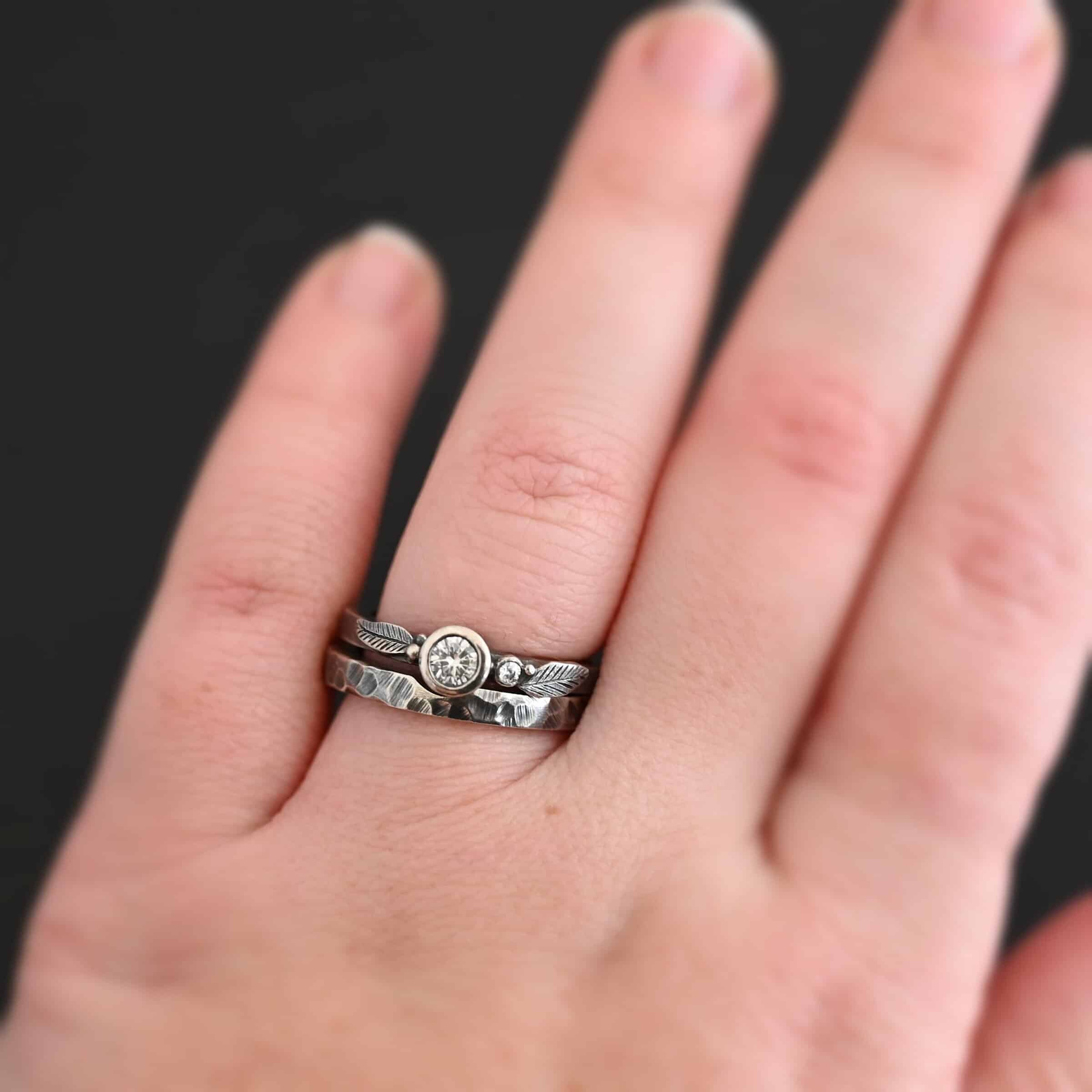 Close up of the "Shining Light" Ring. This ring is handmade out of sterling silver and moissanites. There are two moissanites ~ one as the center stone, and one on the right side of that. The ring has two separate bands that go together, as the top one sits on the bottom one in a groove. There are two silver leaves, two on the top band on each side of the center moissanite. The bottom band is hammered and oxidized silver. There is a model wearing the ring on the engagement ring finger to show how it would look on a hand. The hand is laying against a dark background.