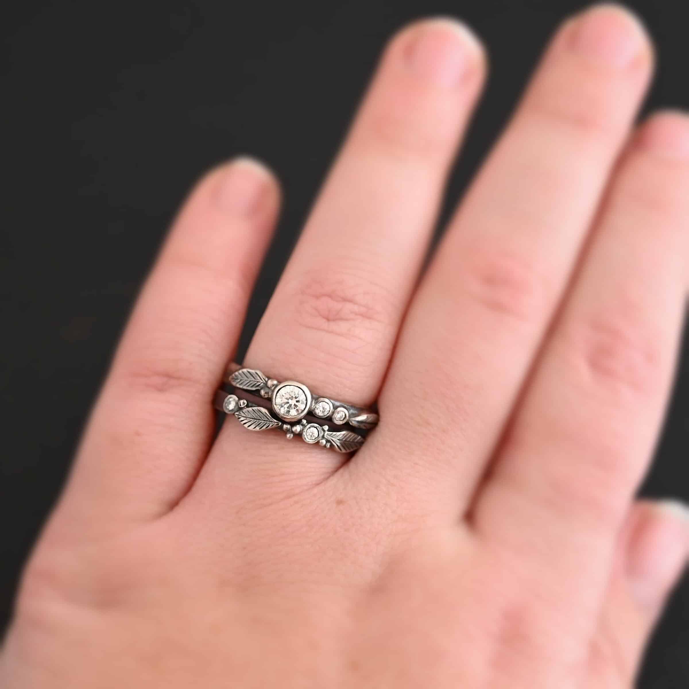 New The Sky Is Everywhere Close up of "The Sky Is Everywhere" Ring. This ring is handmade out of sterling silver and moissanites. There are four moissanites ~ one as the center stone, and three others around the ring. The ring has two separate bands that go together, as the top one sits on the bottom one in a groove. There are four silver leaves, two on each of the bands. There is a model wearing the ring on the engagement ring finger to show how it would look on a hand. The hand is laying against a dark background.