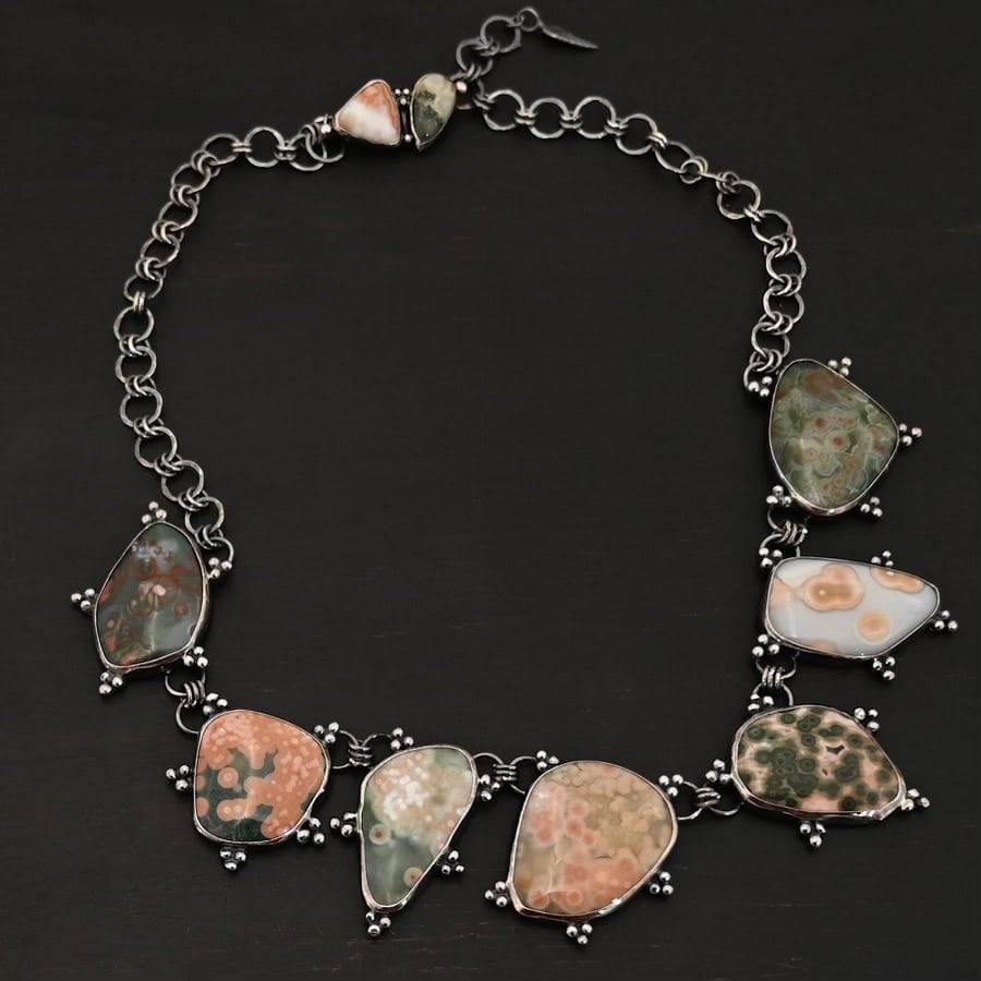 Look For Adventure Necklace The "Look For Adventure" Necklace. Made out of sterling silver and ocean jasper. There are nine ocean jasper stones that create this necklace. Seven of them are large stones, and there are two smaller ones on the back that are a part of the clasp for the necklace. Each stone is a various shape, size, and color from one another. There is a lot of green and orange colors, with a bit of red in some of the stones. Each ocean jasper has multiple sterling silver dots going around the stone setting. The chain is made out of sterling silver hammered circular links that are oxidized. Necklace is sitting against a dark background.