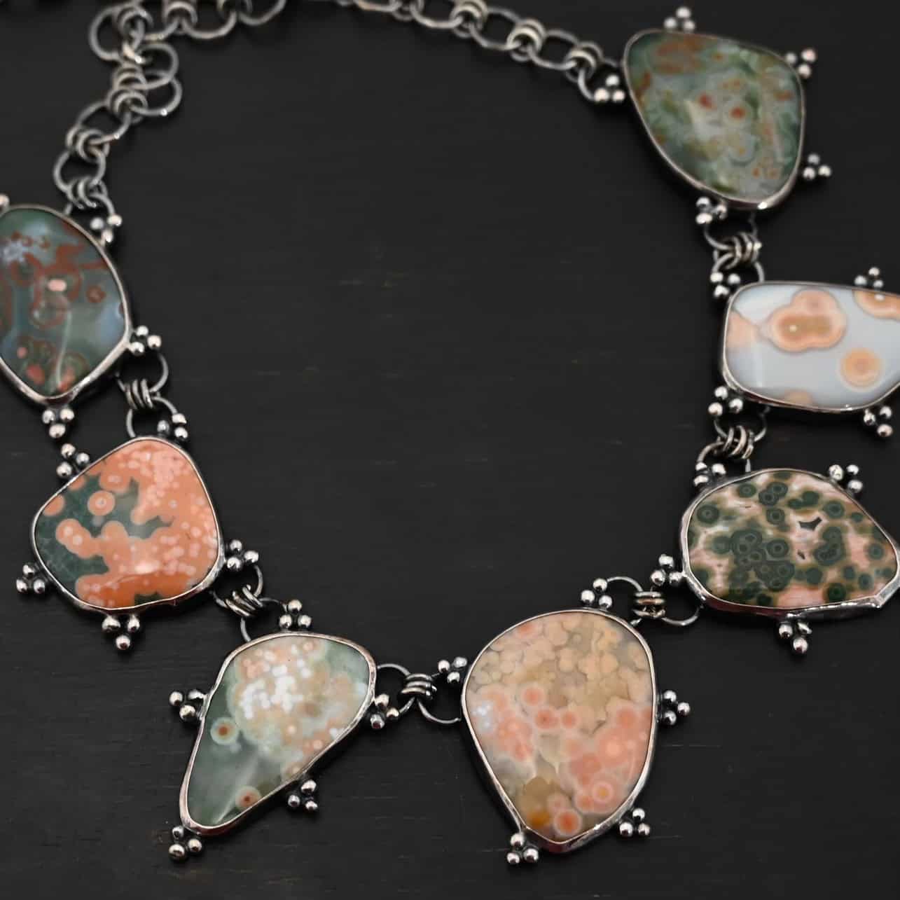 Look For Adventure Necklace The "Look For Adventure" Necklace. Made out of sterling silver and ocean jasper. There are nine ocean jasper stones that create this necklace. Seven of them are large stones, and there are two smaller ones on the back that are a part of the clasp for the necklace. Each stone is a various shape, size, and color from one another. There is a lot of green and orange colors, with a bit of red in some of the stones. Each ocean jasper has multiple sterling silver dots going around the stone setting. The chain is made out of sterling silver hammered circular links that are oxidized. Necklace is sitting against a dark background.