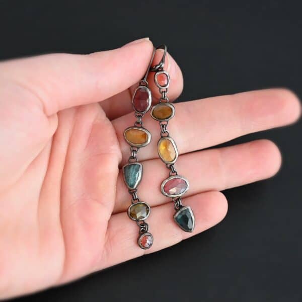 "Living In Color" Earrings as shown up close. Made out of sterling silver and ten sapphires with leverback ear wires. The sapphires are multiple colors. Earrings are set against the fingertips of a hand.