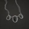 Close up of the "Escape To Tranquility" Necklace. Made out of sterling silver and three black onyx. The middle black onyx is the largest, with two smaller ones on the side. Each black onyx is set in silver and has silver balls going around it. Each black onyx is connected with two jump rings. Necklace is set against a dark background. Each side of the necklace is connected to two chains. Necklace is set against a dark background.