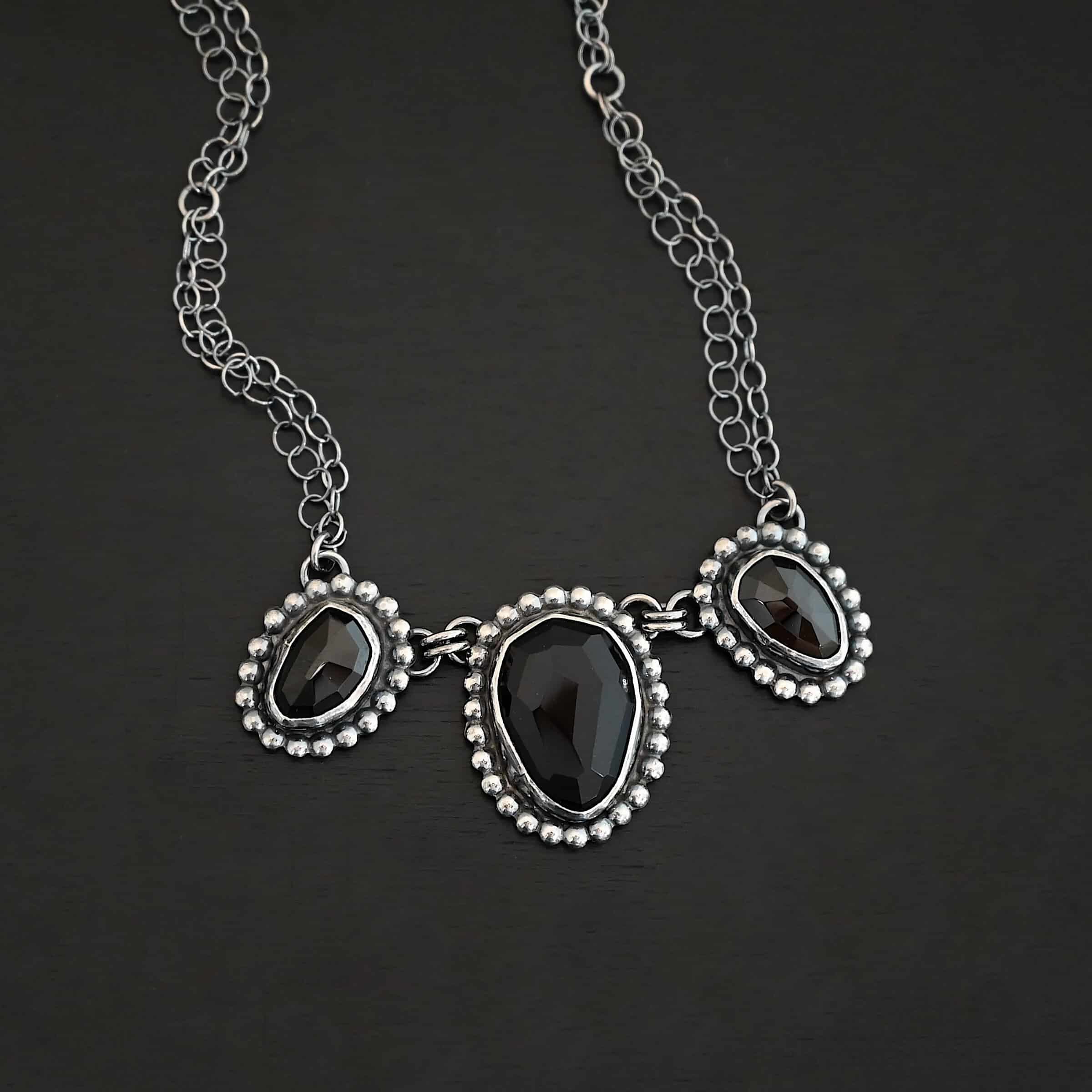 Close up of the "Escape To Tranquility" Necklace. Made out of sterling silver and three black onyx. The middle black onyx is the largest, with two smaller ones on the side. Each black onyx is set in silver and has silver balls going around it. Each black onyx is connected with two jump rings. Necklace is set against a dark background. Each side of the necklace is connected to two chains. Necklace is set against a dark background.