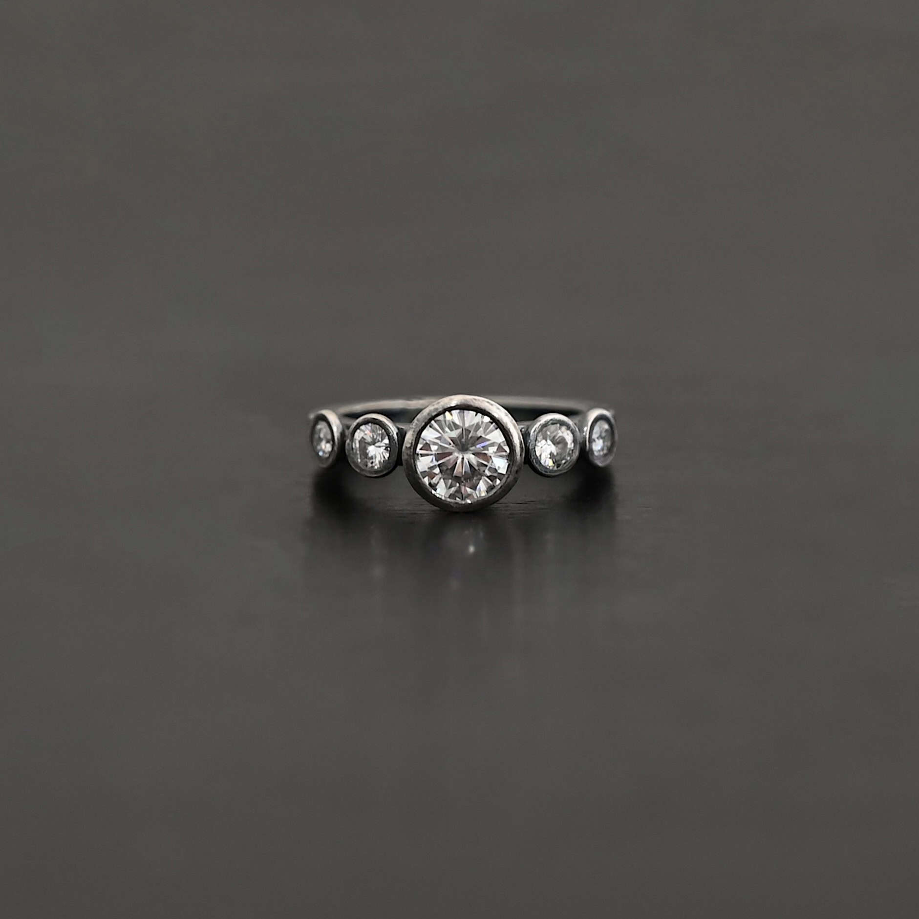 Heart Of Silver Ring New Photo of the "Heart Of Silver" Ring. There is a large moissanite in the center of the ring, with two smaller moissanites on each side of it, making for five total moissanites on the ring. Ring is set against a dark background.
