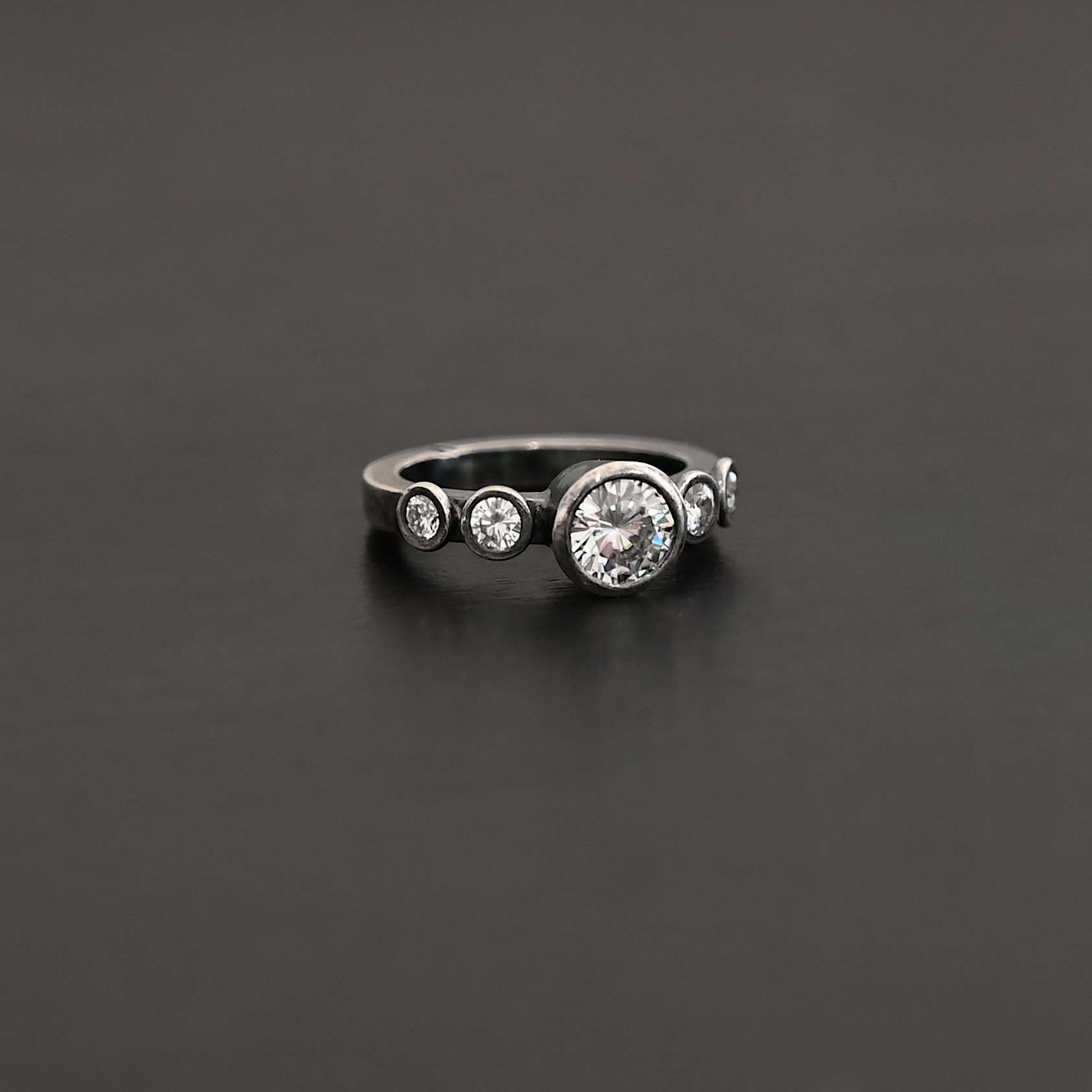 Heart Of Silver Ring New Photo of the "Heart Of Silver" Ring. There is a large moissanite in the center of the ring, with two smaller moissanites on each side of it, making for five total moissanites on the ring. Ring is set against a dark background.
