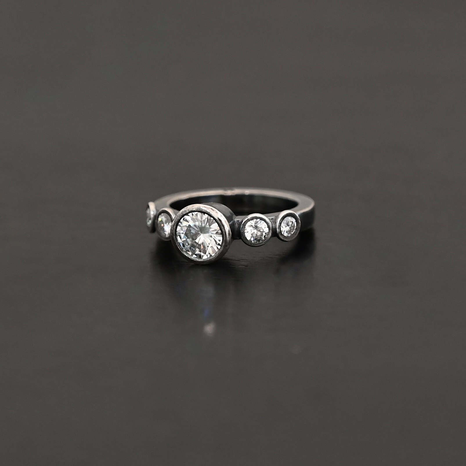 Heart Of Silver Ring New Photo of the "Heart Of Silver" Ring. There is a large moissanite in the center of the ring, with two smaller moissanites on each side of it, making for five total moissanites on the ring. Ring is set against a dark background.