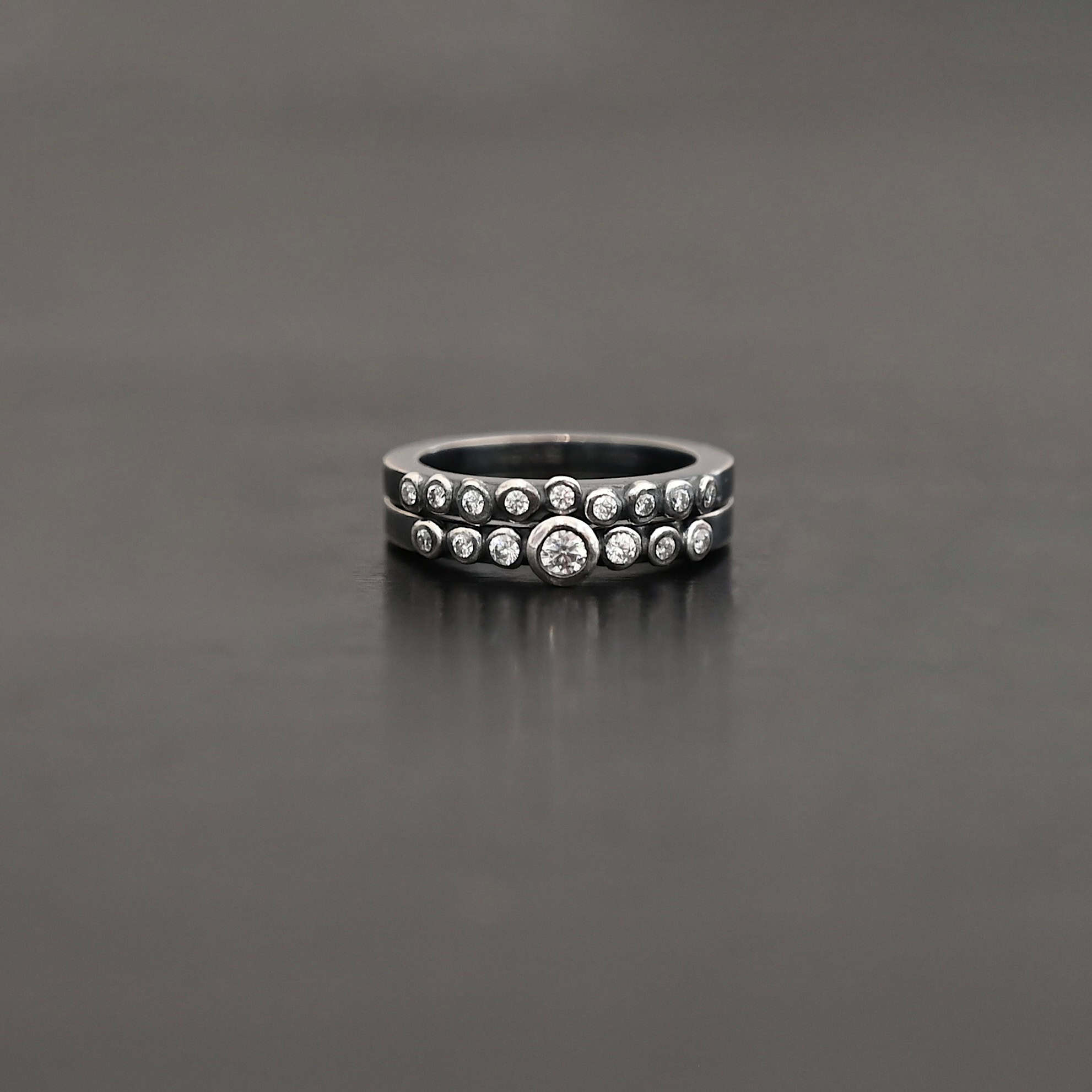 "Aura Chic" Ring as shown up close. Made out of 16 moissanites. There are two bands. The top band contains 7 moissanites. It also contains the largest moissanite, with three on the left side of that and three on the right. The bottom band contains 9 moissanites, all the same size. Ring is set against a dark background.
