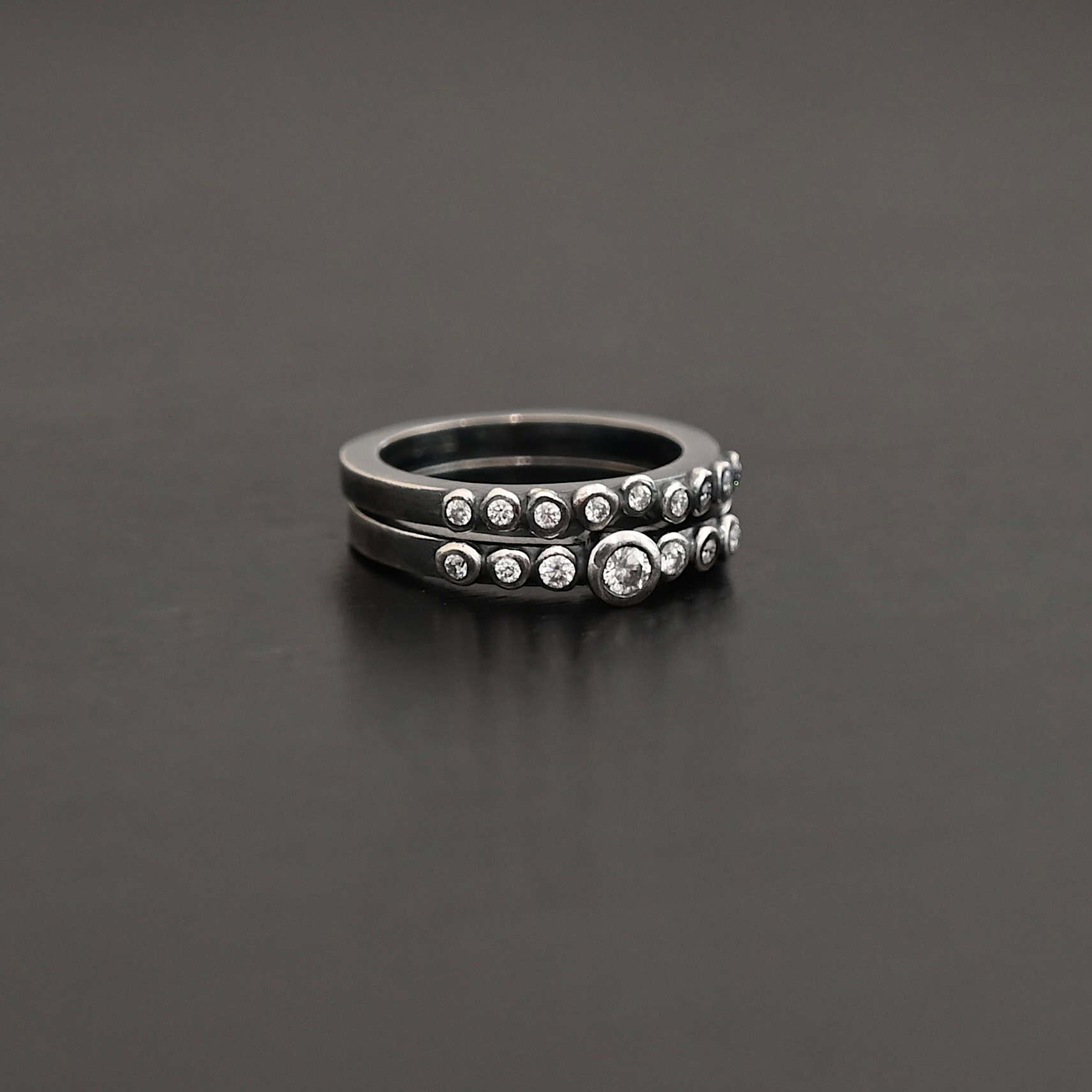 "Aura Chic" Ring as shown up close. Made out of 16 moissanites. There are two bands. The top band contains 7 moissanites. It also contains the largest moissanite, with three on the left side of that and three on the right. The bottom band contains 9 moissanites, all the same size. Ring is set against a dark background.