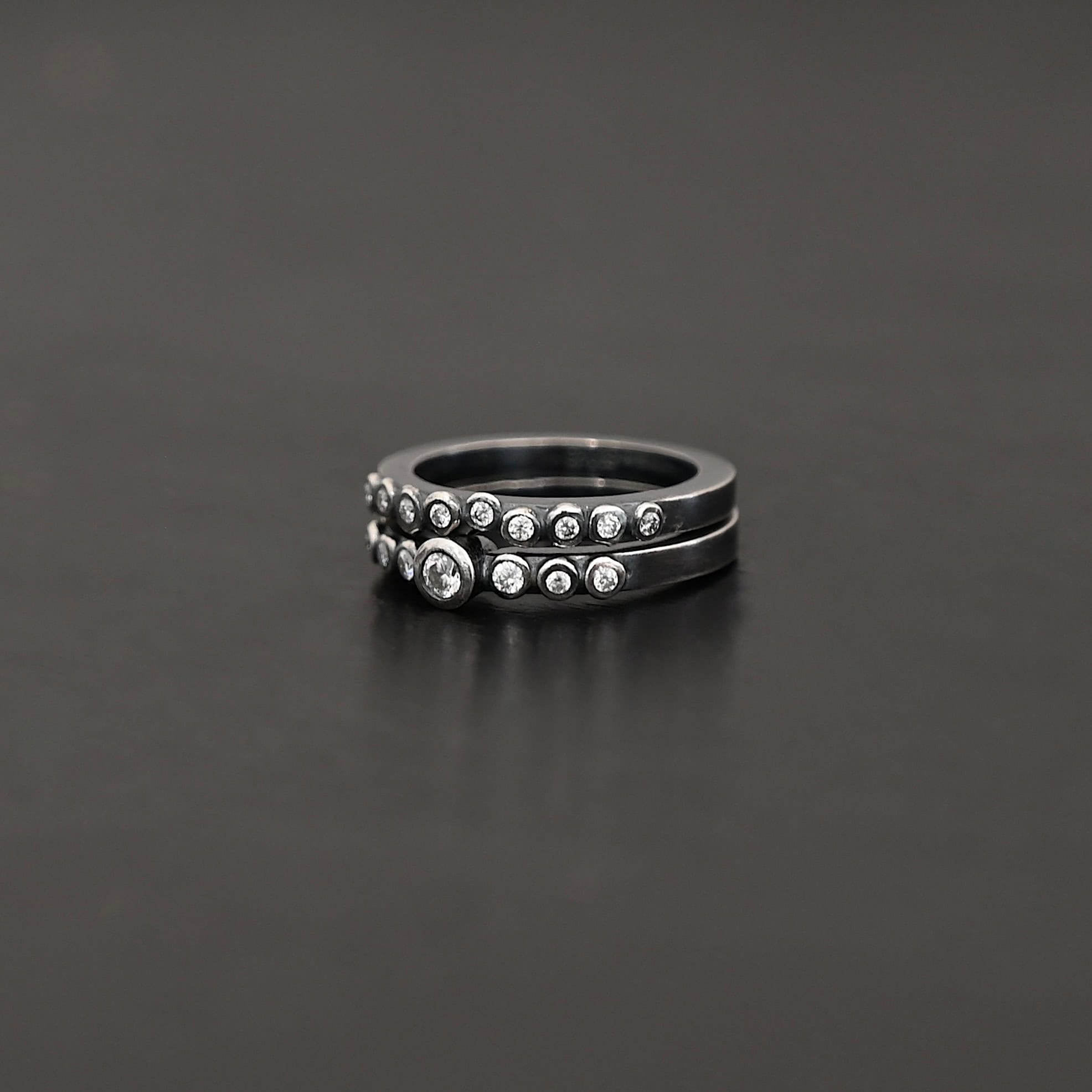 "Aura Chic" Ring as shown up close. Made out of 16 moissanites. There are two bands. The top band contains 7 moissanites. It also contains the largest moissanite, with three on the left side of that and three on the right. The bottom band contains 9 moissanites, all the same size. Ring is set against a dark background.