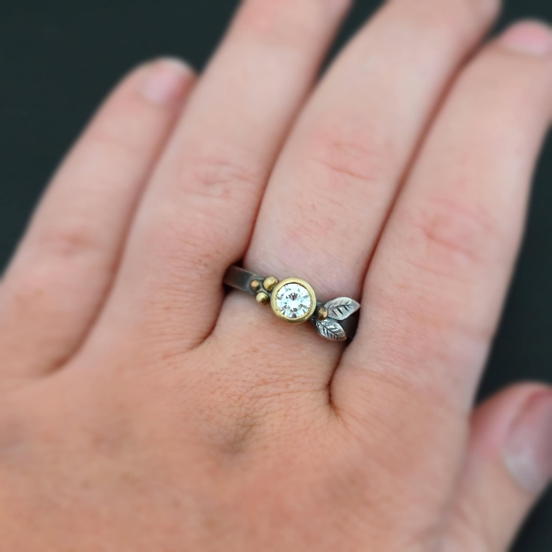 Here's To Forever Ring "Here's To Forever" Ring as shown up close. Ring is made out of sterling silver, gold, and moissanite. There are two sterling silver engraved leaves on the left side of the ring with a gold ball at the end of them, and closest to the moissanite. There are three gold balls on the right side of the ring. In the center of the ring is a moissanite that is set in gold. Ring is worn by a model on their engagement ring finger to demonstrate size and width of the ring.