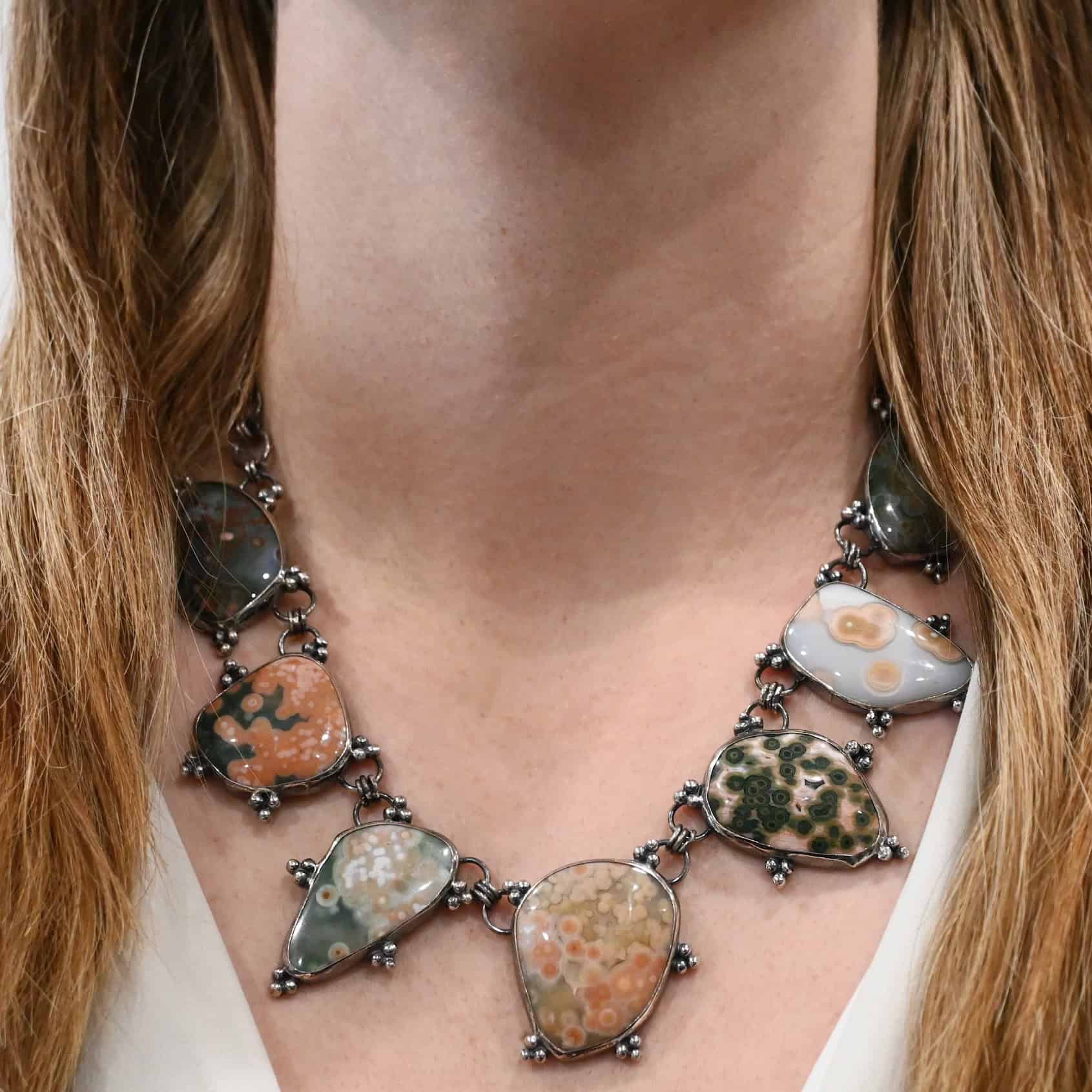 Look For Adventure Necklace The "Look For Adventure" necklace as shown on a model. Necklace is made out of sterling silver and ocean jasper. Necklace has separate ocean jasper pieces that are connected by large, hammered, oxidized links that are circles. The ocean jasper pieces are a variety of colors, but they all have a lot of dots. Model is wearing a white v-neck shirt.